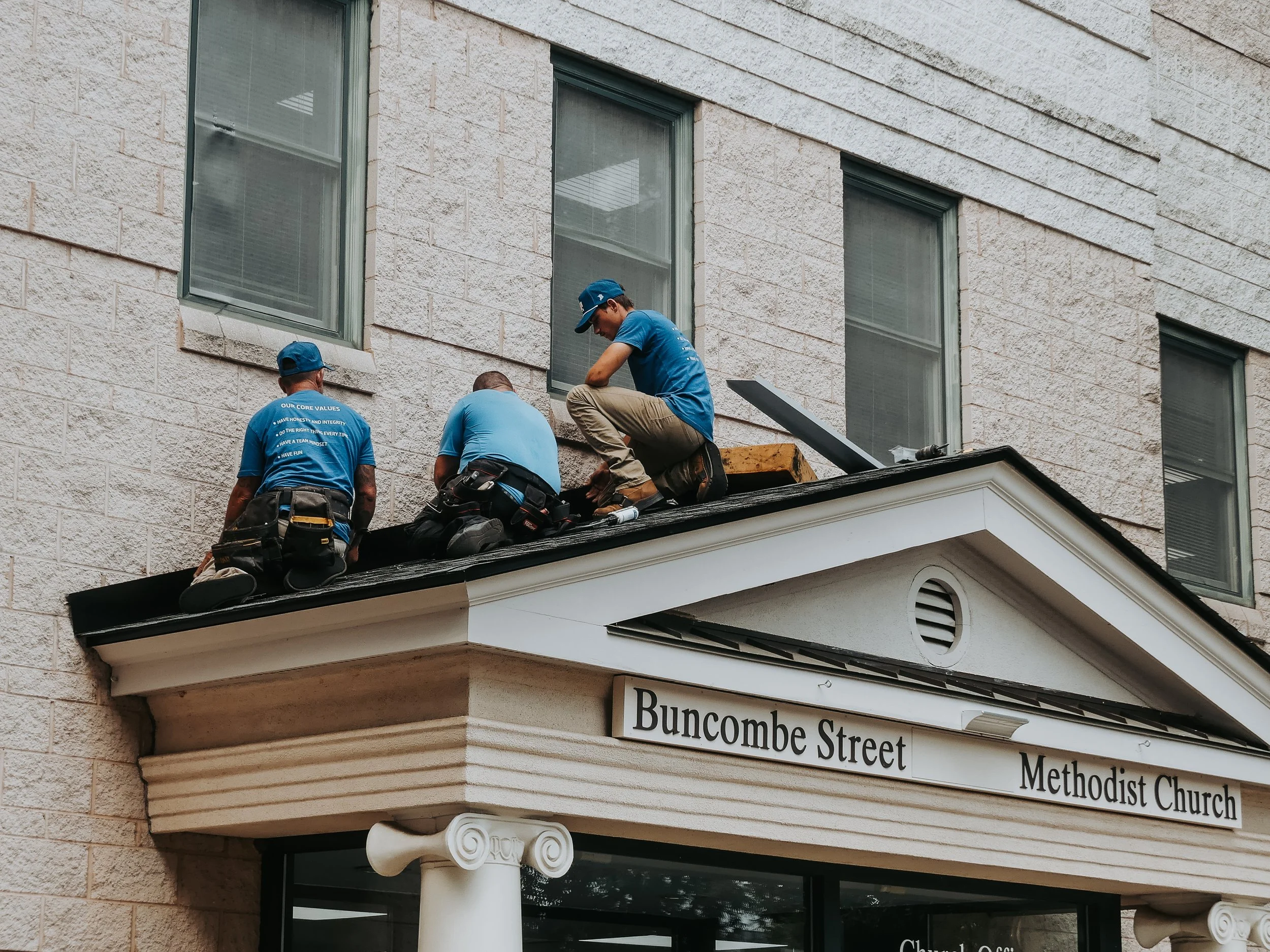 Three workers in blue shirts and caps on the roof of a Methodist church, installing or repairing the roof.