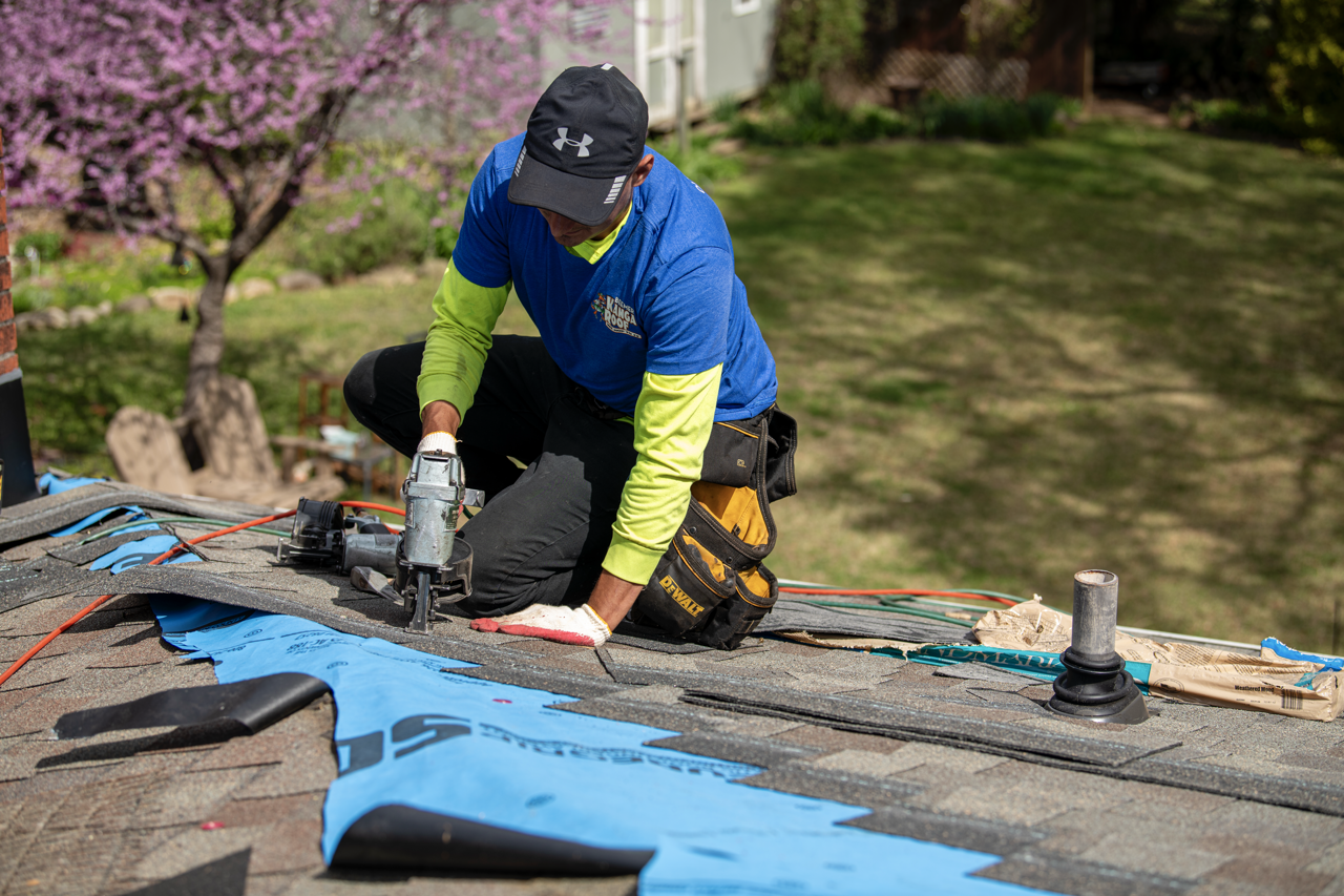 A man wearing a blue long sleeve shirt, black cap, and black work belt kneeling on a roof, using a power tool to perform roofing work, with roofing materials and tools nearby.
