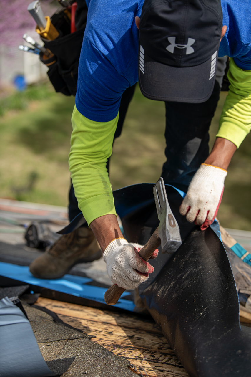 A person wearing work gloves, a black cap, and outdoor clothing working with a hammer on a roofing project outdoors.