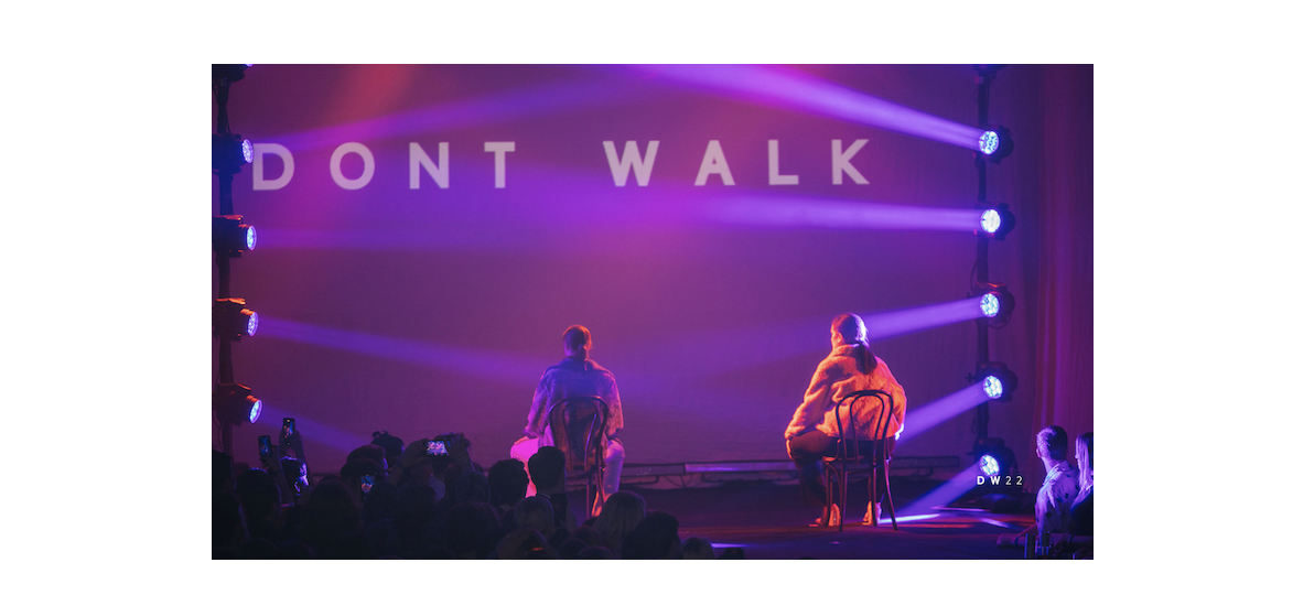 Two people sitting on chairs on stage with purple and pink lighting, large screen behind them displaying the words "DONT WALK." Audience watching and taking photos.