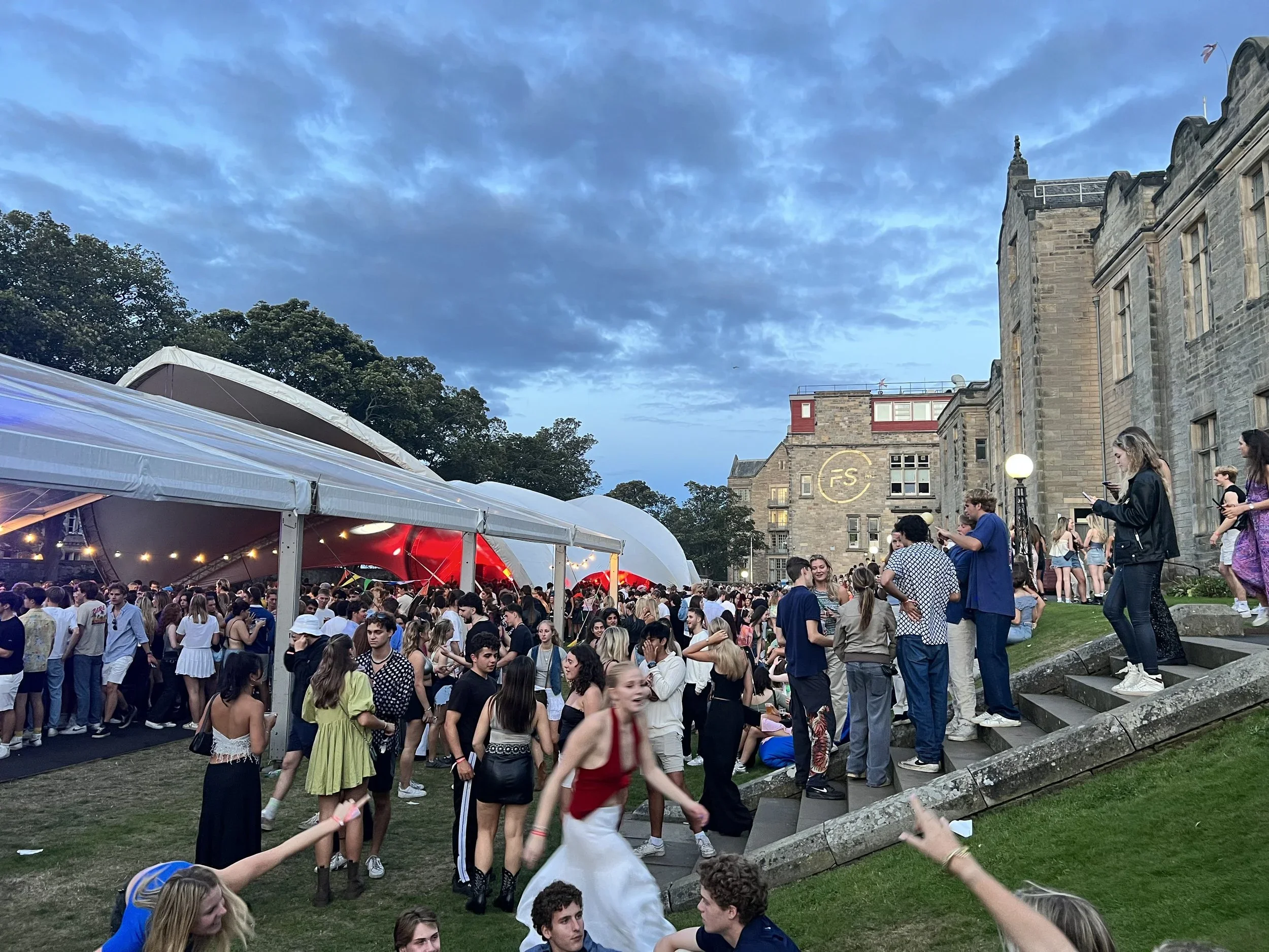 A large outdoor gathering of people, mostly young adults, by a historic stone building with a lit circular logo on it. There is a white tent with string lights on the left side of the image. The sky is partly cloudy with a hint of evening light.