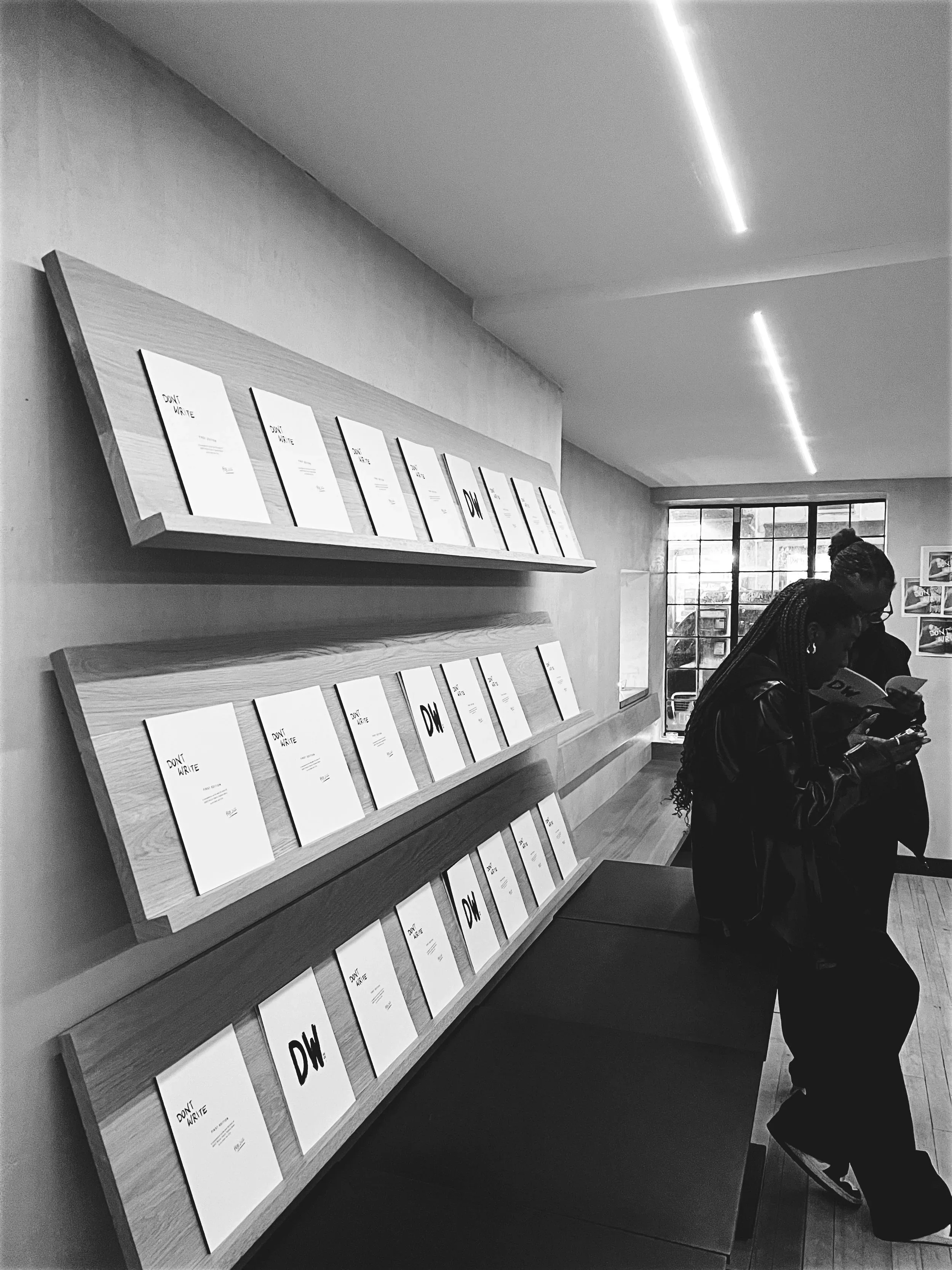 Two women reading books or magazines in a modern interior space with shelves displaying identical publications titled 'DON'T WRITE' and 'DW'.
