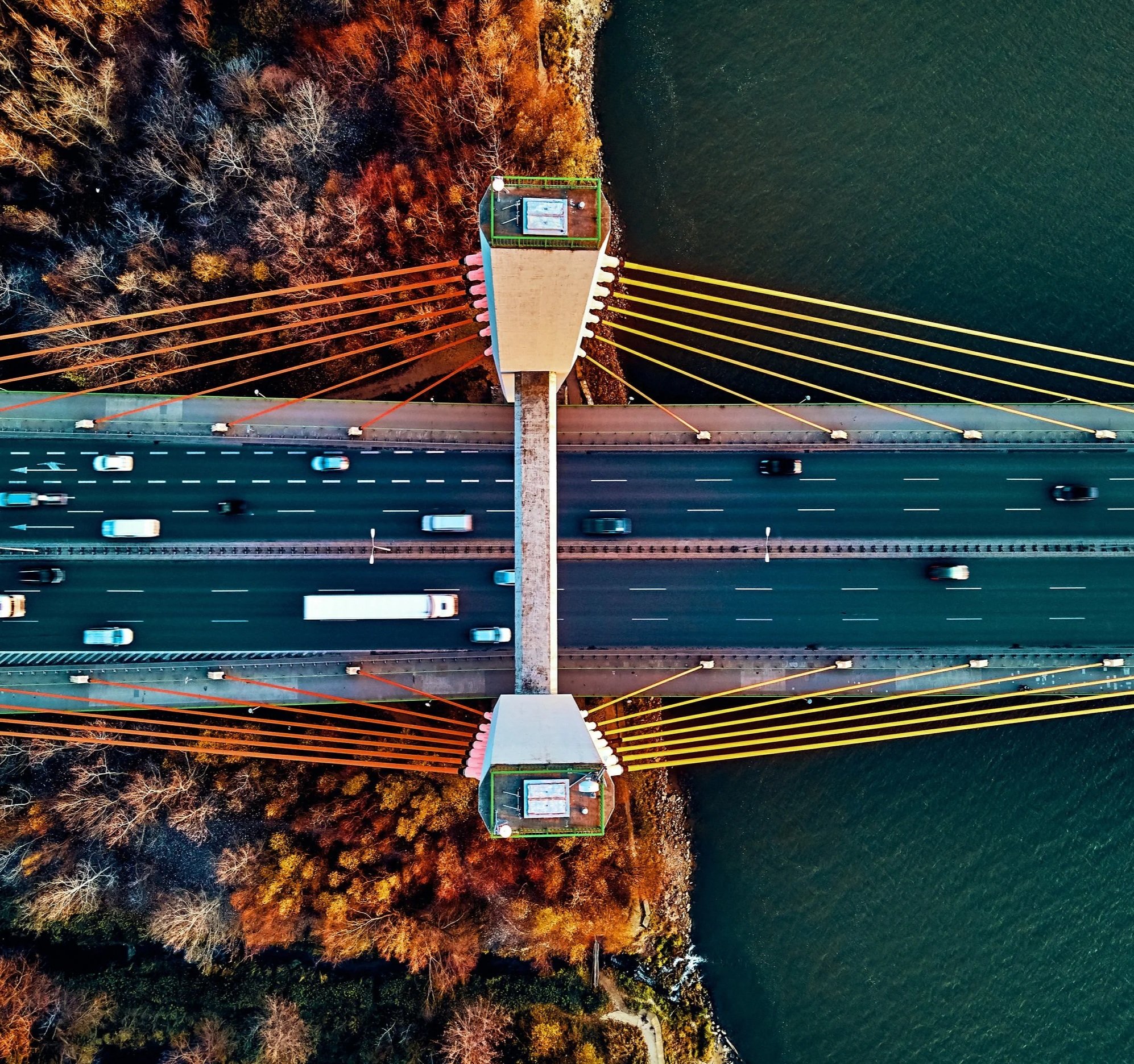 An aerial view of a bridge spanning across a body of water, with a paved walkway and vehicular lanes, surrounded by trees with autumn foliage.