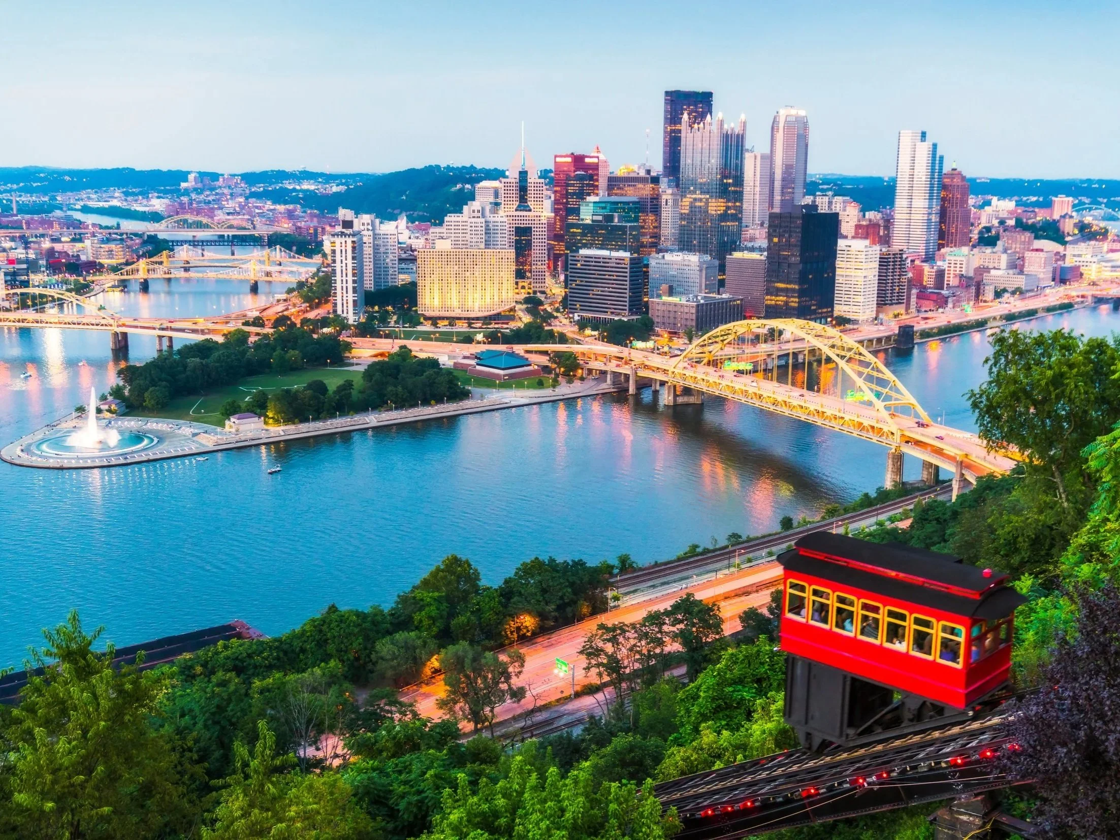 A city skyline at dusk with tall skyscrapers, bridges over a river, and a double-decker trolley in the foreground on a hill surrounded by lush green trees.