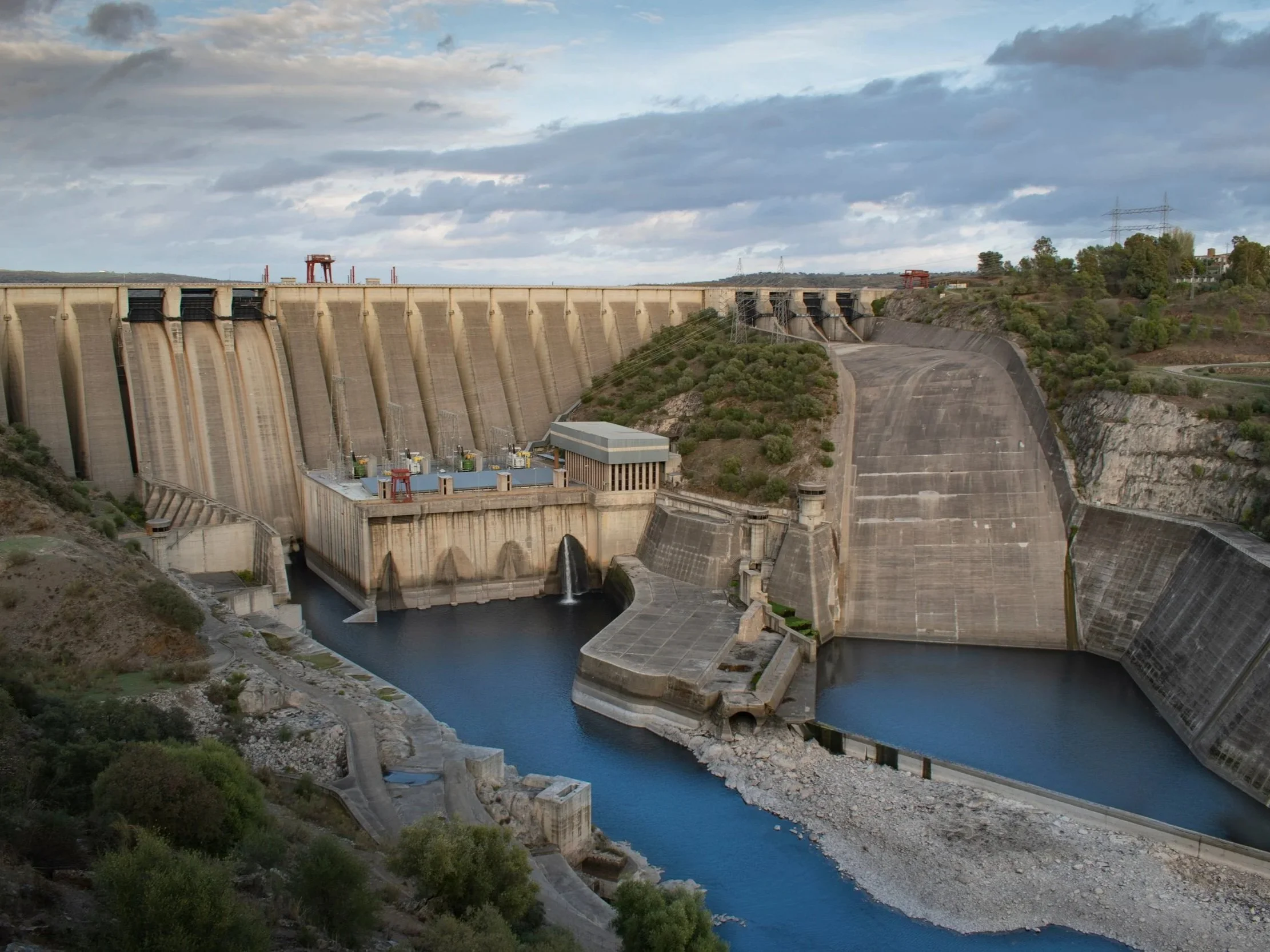 A large dam with concrete spillways releasing water, situated in a hilly landscape with some trees and buildings on the hillside, under a cloudy sky.