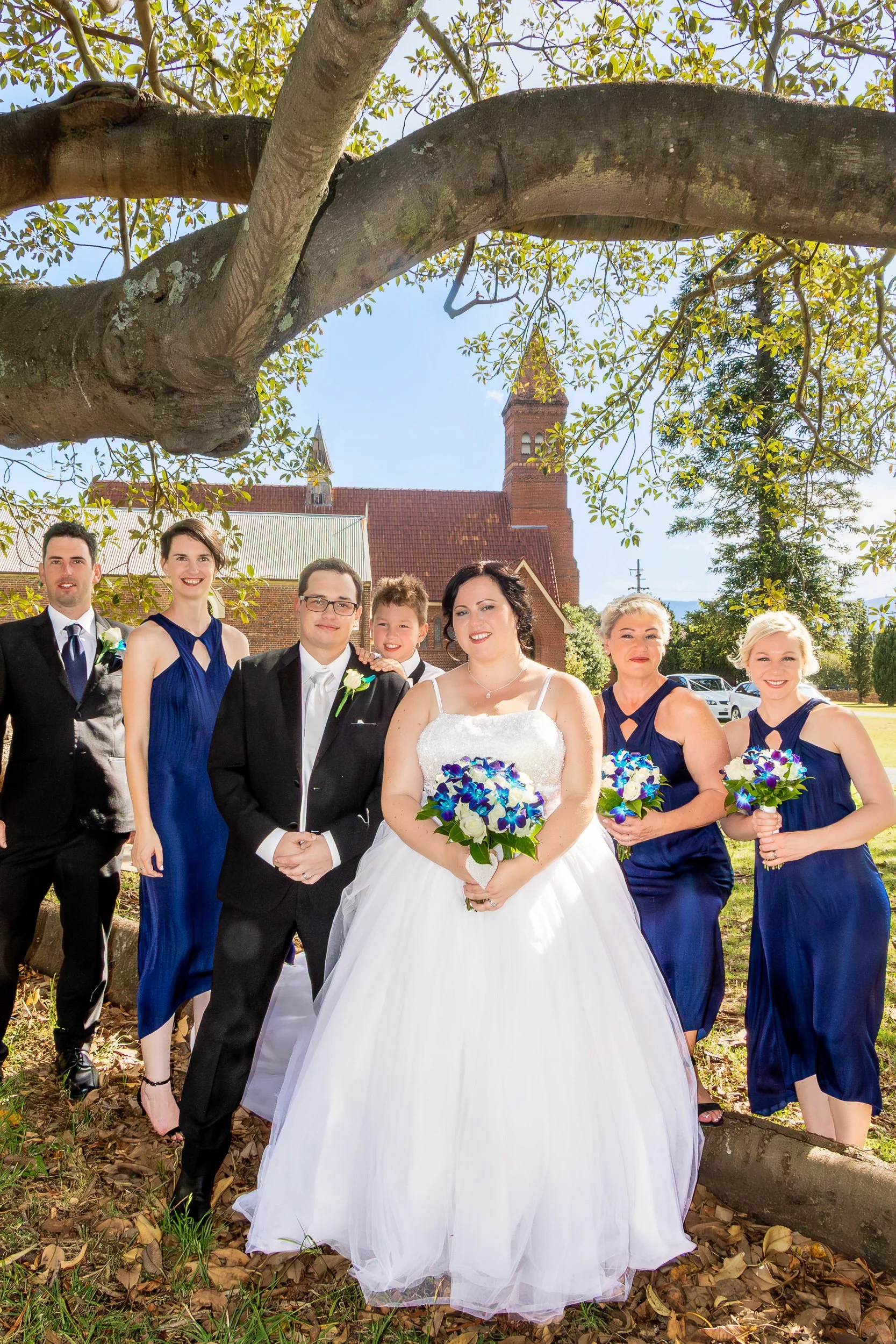  Bride, groom and wedding party gathered beneath a large tree. 