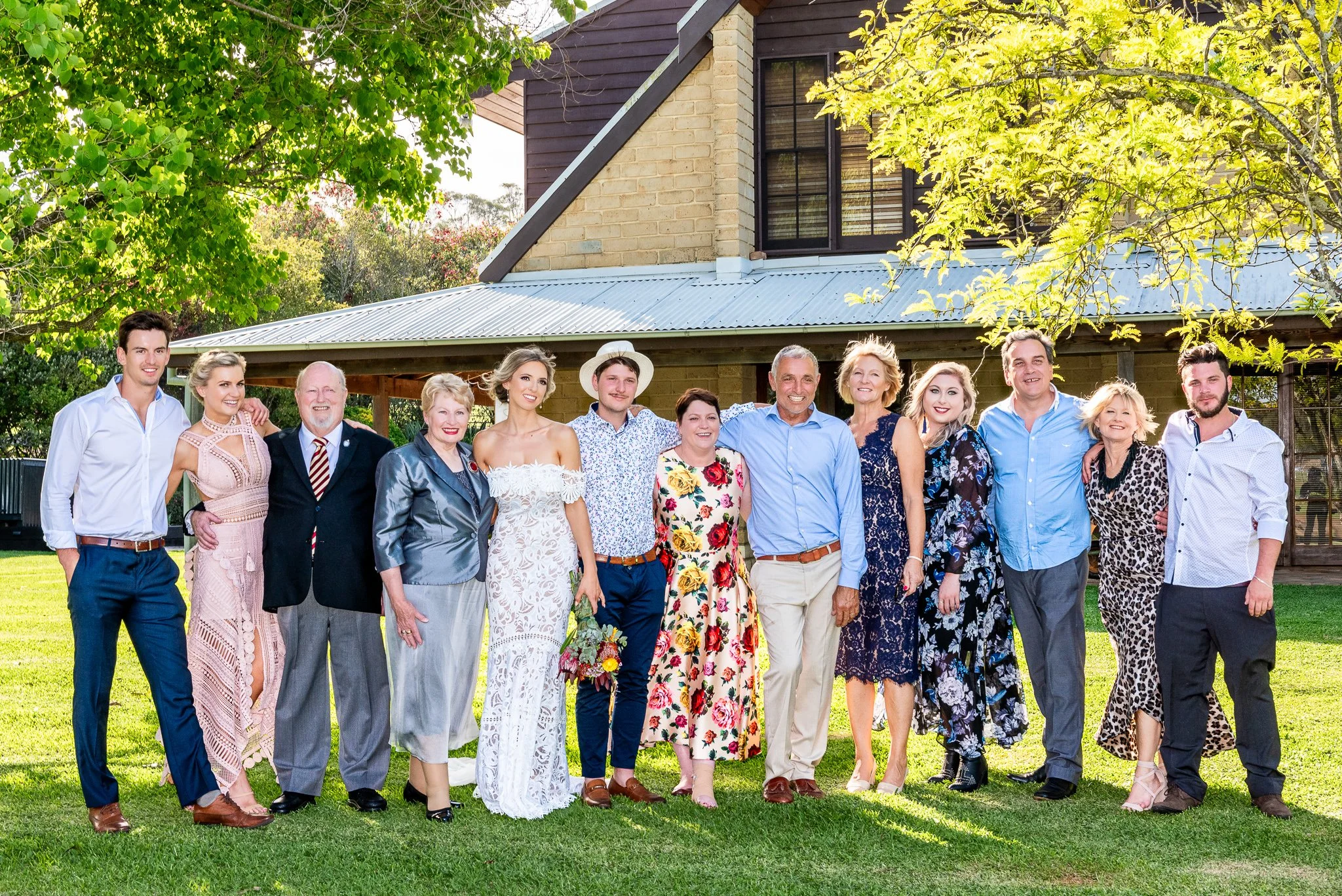 Bride and groom standing with family members for a formal outdoor wedding group portrait in front of a country-style building.