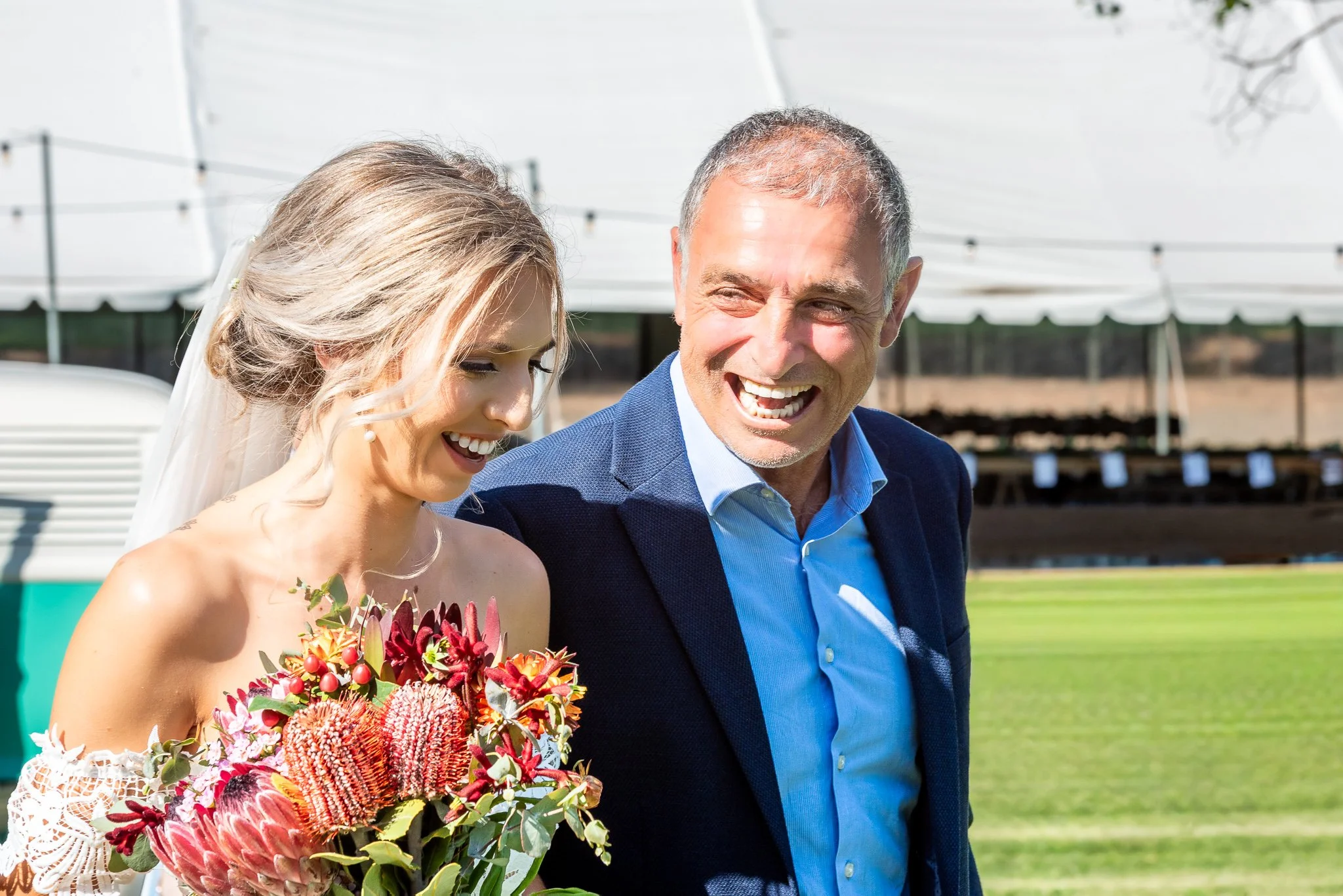 Bride smiling while walking outdoors with a family member and holding a native flower bouquet on her wedding day.