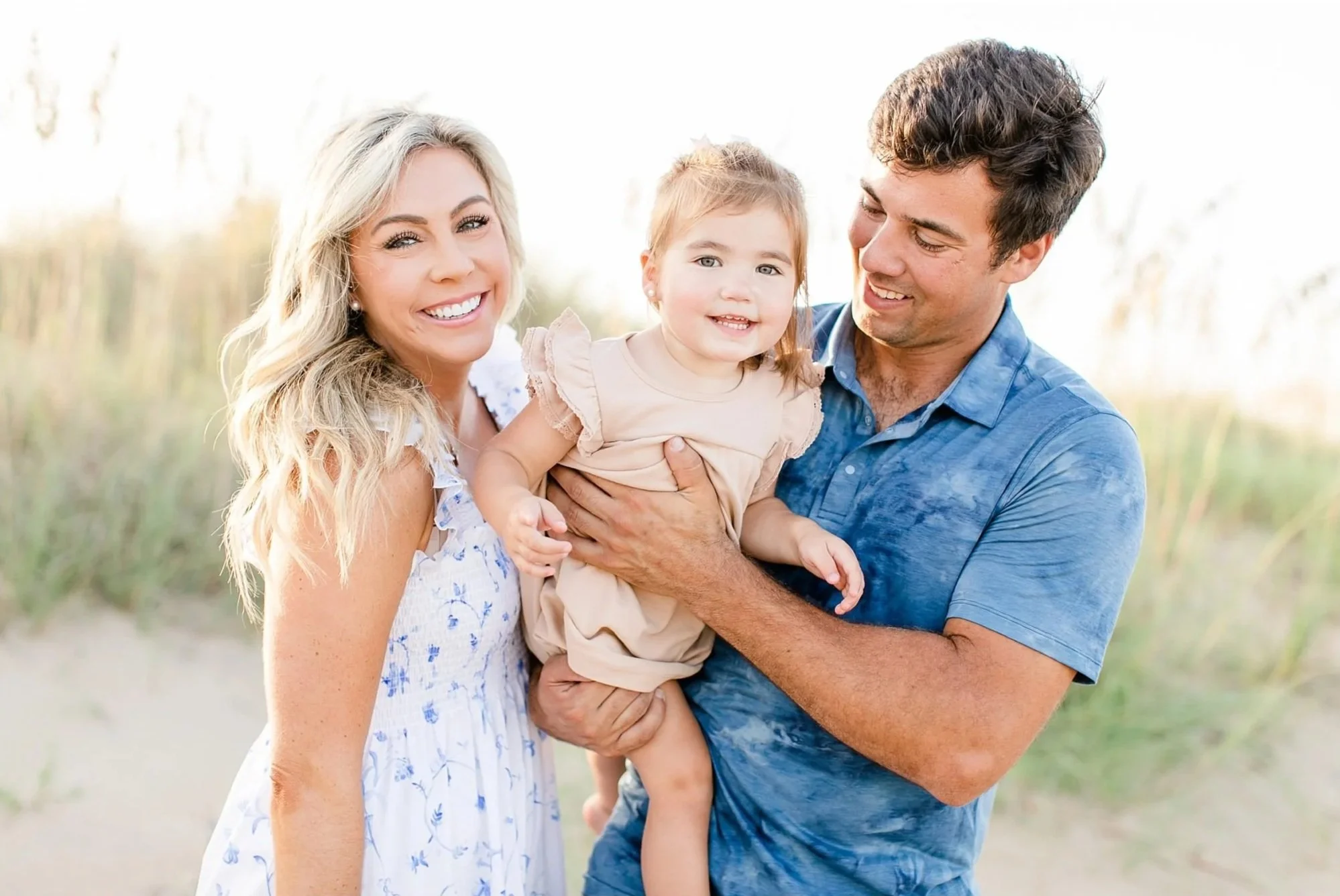 Parents holding their young child during a bright outdoor family portrait near the beach.