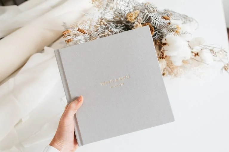 Wedding photo album held beside dried flowers and soft fabric, showing a finished printed keepsake.