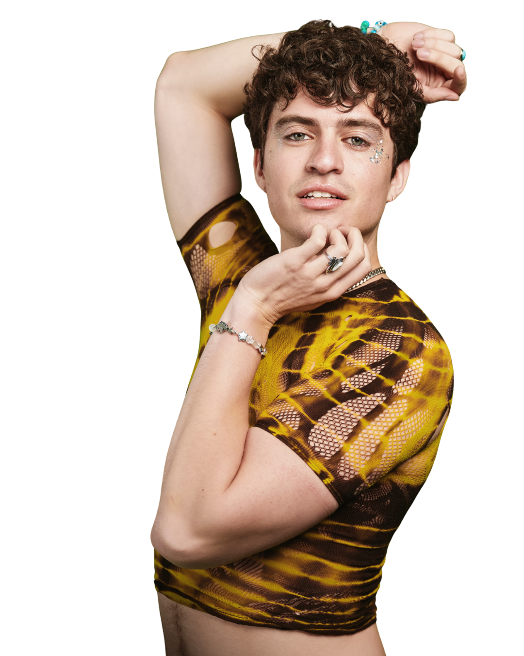 Person with curly hair wearing a yellow and black patterned mesh top posing with one arm raised and hand touching their face, wearing jewelry and glitter on their face, against a dark background.