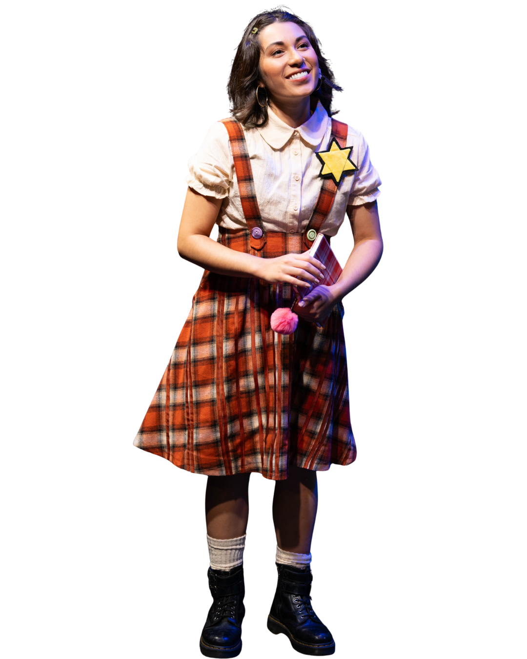 Young woman wearing a plaid dress and plaid suspenders, holding a small folder, with a star-shaped badge on her shirt, standing on a stage with black background.