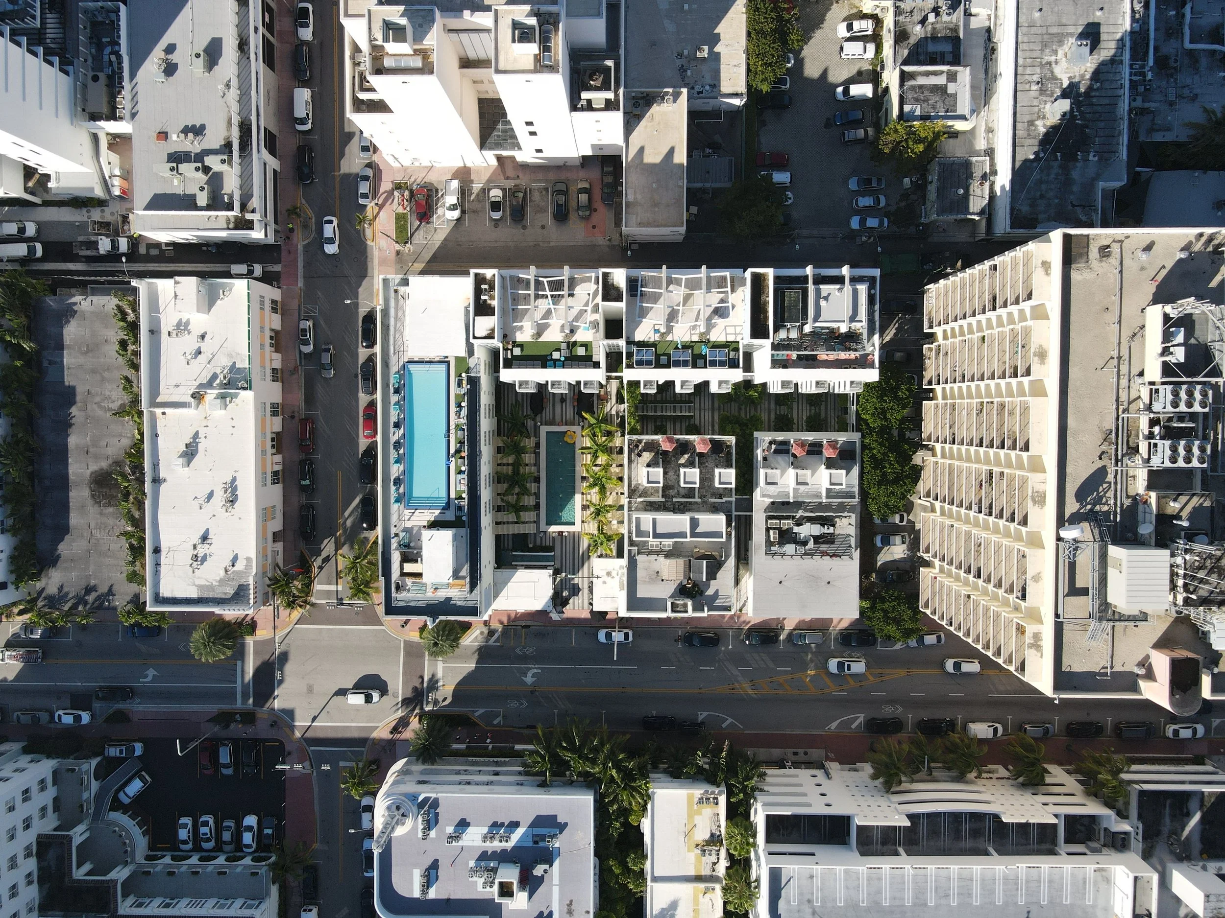 Aerial view of a city block showing multi-story buildings, parking lots, streets, and some trees, with shadows indicating a sunny day.