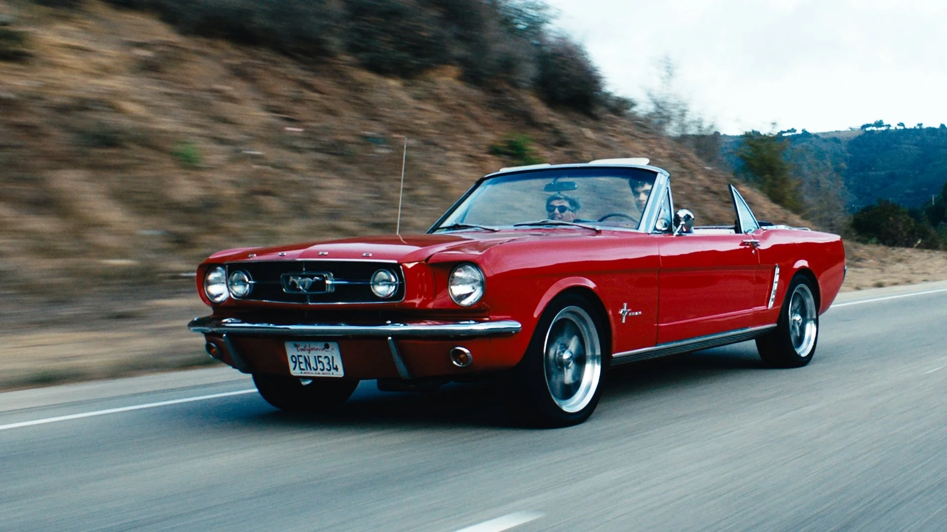A red vintage Ford Mustang convertible driving on a highway with a hillside in the background.