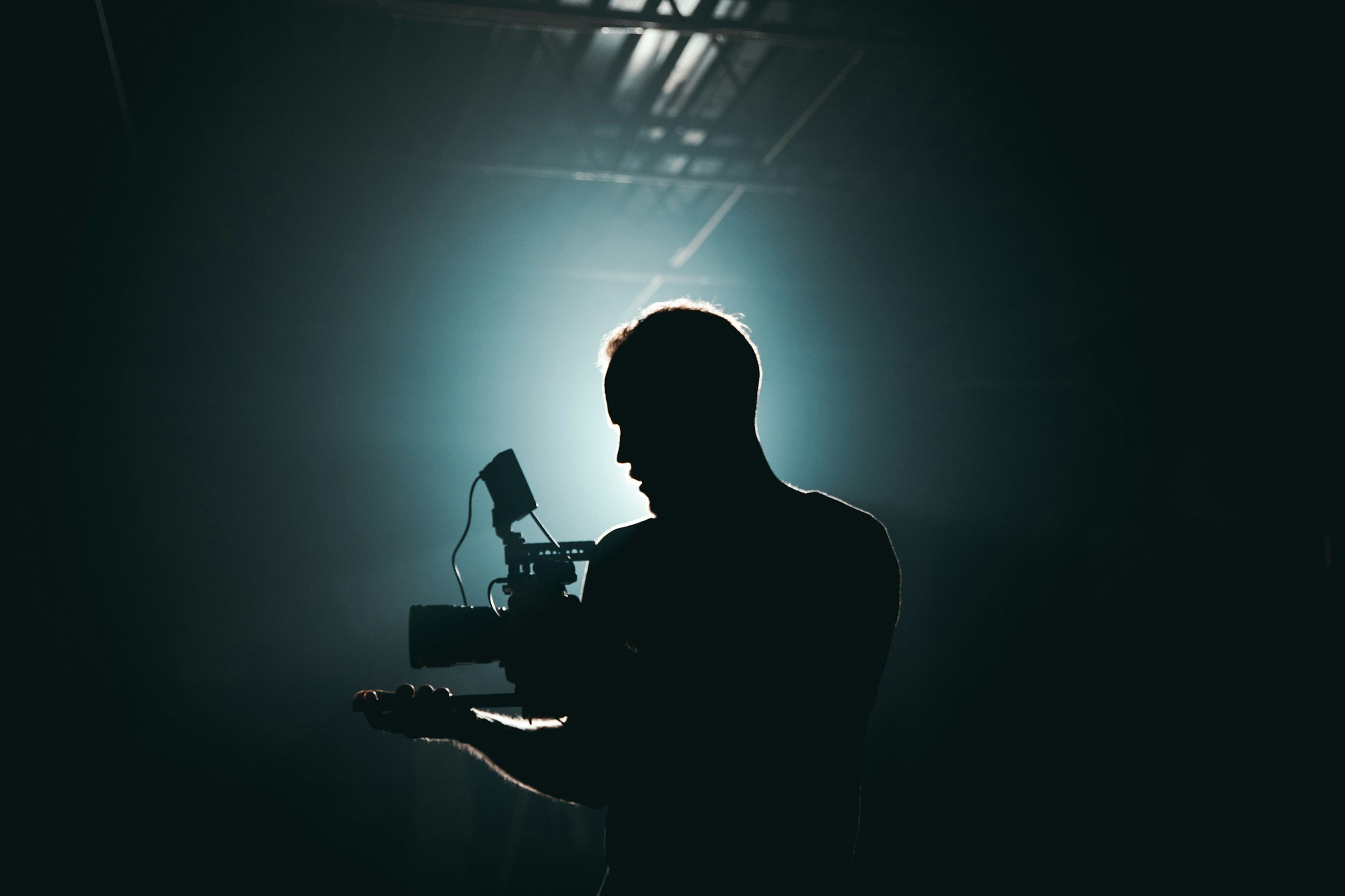 Silhouette of a person holding a professional video camera against a backlit dark background.