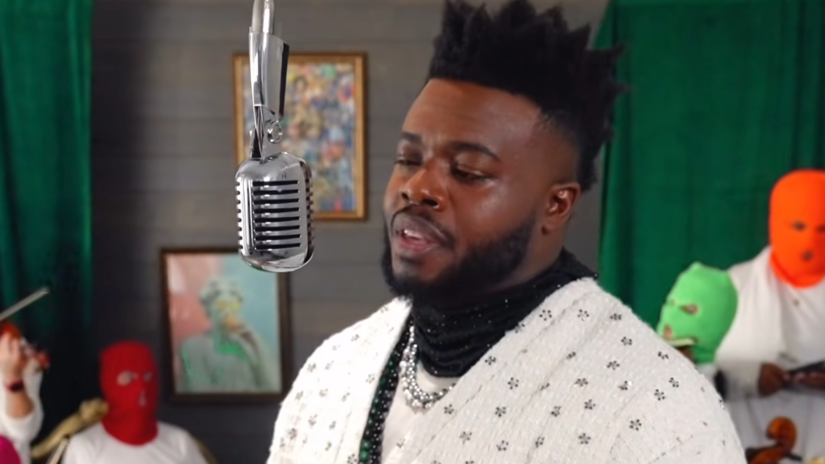 A man with styled hair and beard singing into a vintage microphone with several masked individuals in the background, some holding instruments, in a room with green curtains and framed artwork.