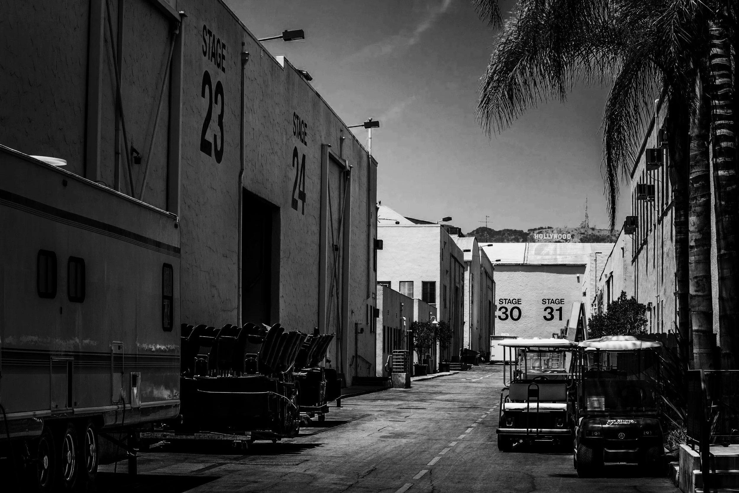 Backstage area with stage numbers 23, 24, 30, and 31 visible on buildings, parking lot with equipment, trees, and the Hollywood sign in the distance.