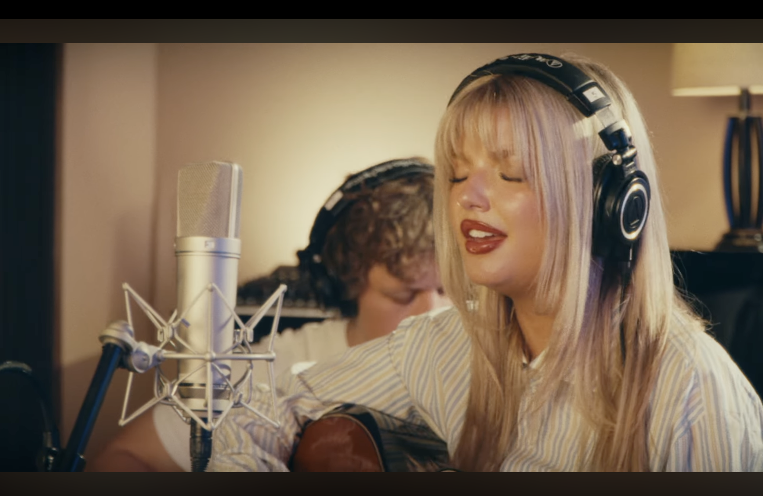 A woman with blonde hair wearing headphones singing into a microphone while playing guitar in a recording studio, with a man in the background playing guitar.