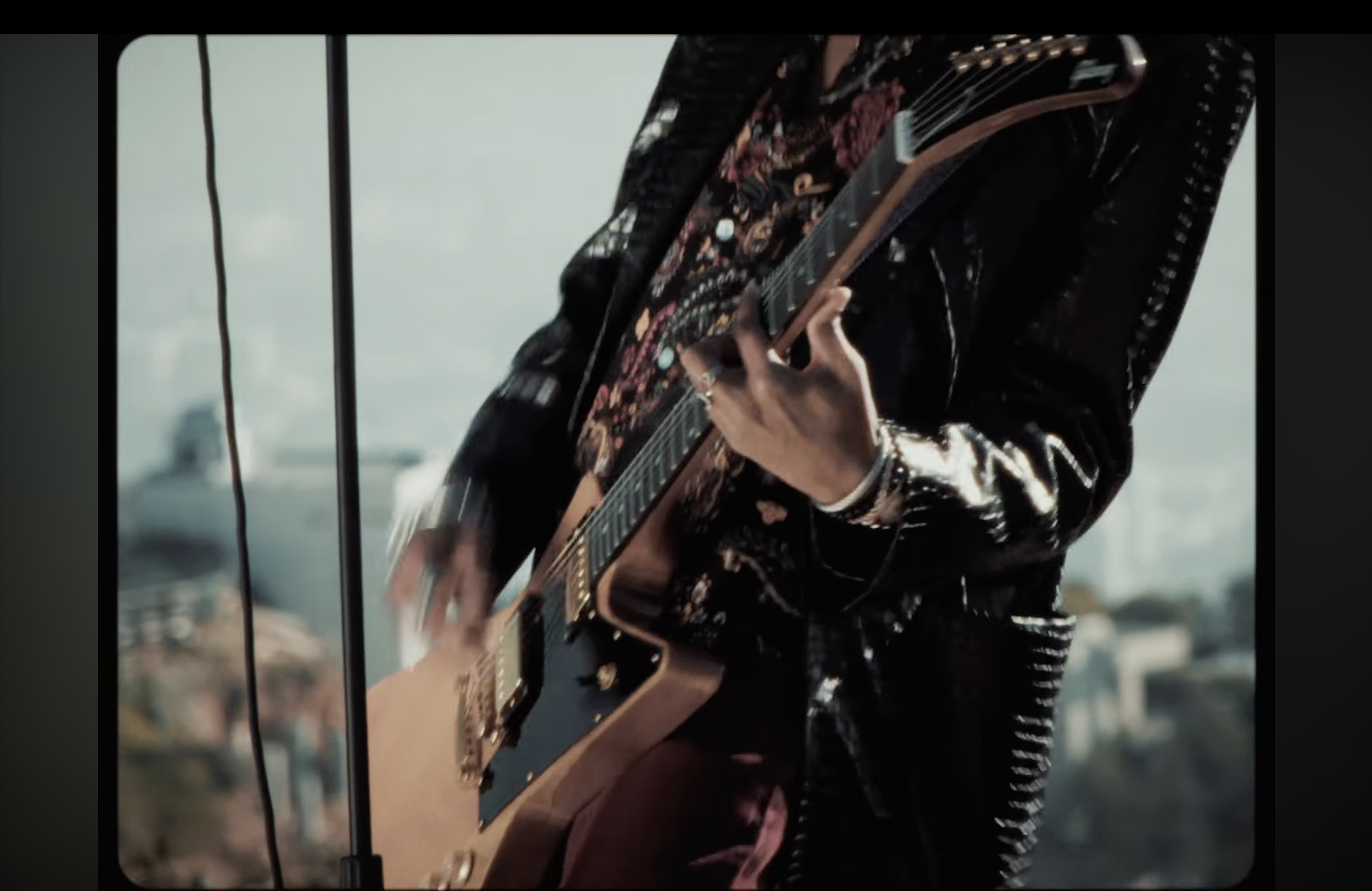Person playing an electric guitar, wearing a black leather jacket, with cityscape and water in the background.