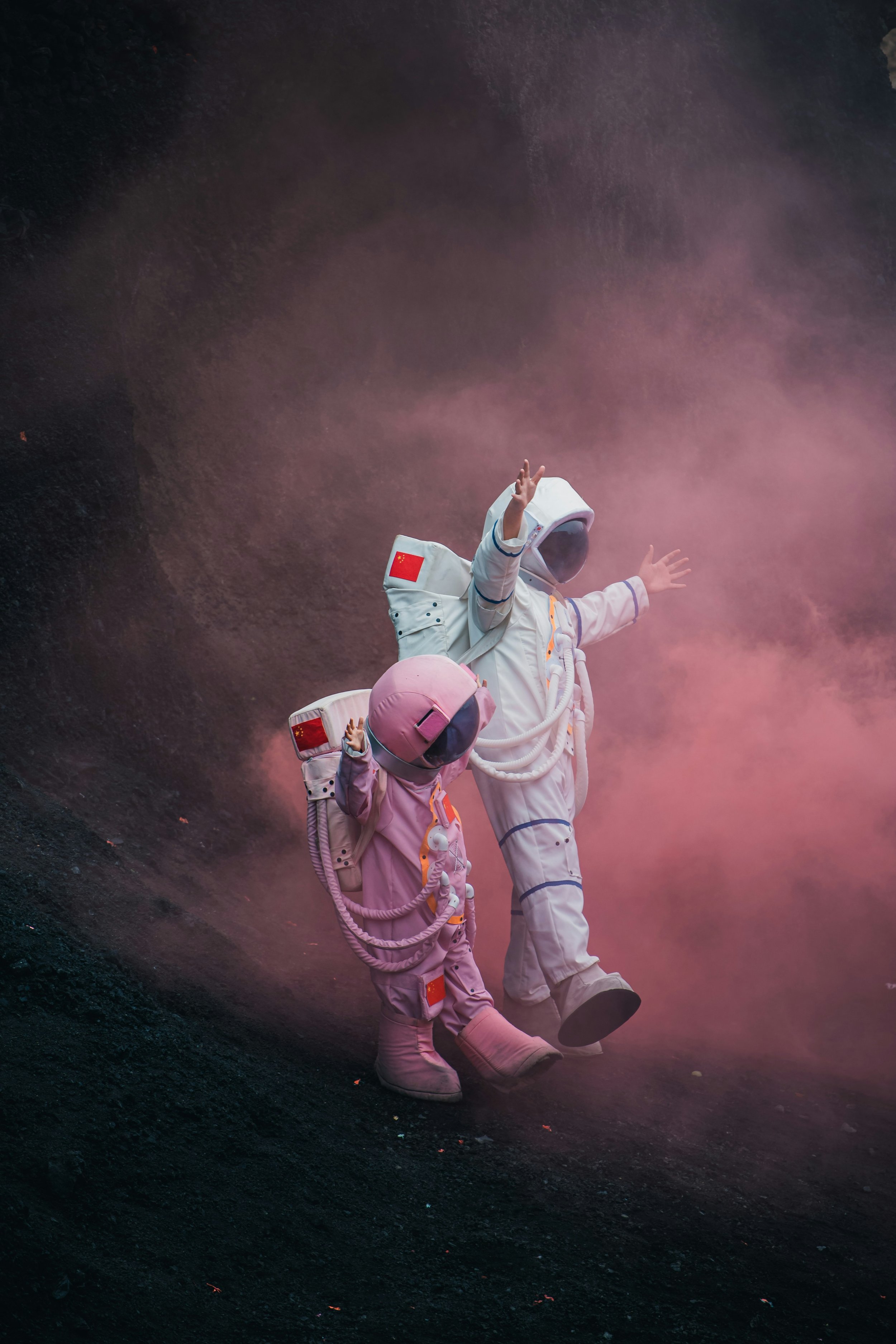 Two astronauts, one adult and one child, walking on a dark terrain with pinkish smoke around them, dressed in suits with Chinese flags on the shoulders, raising their arms.