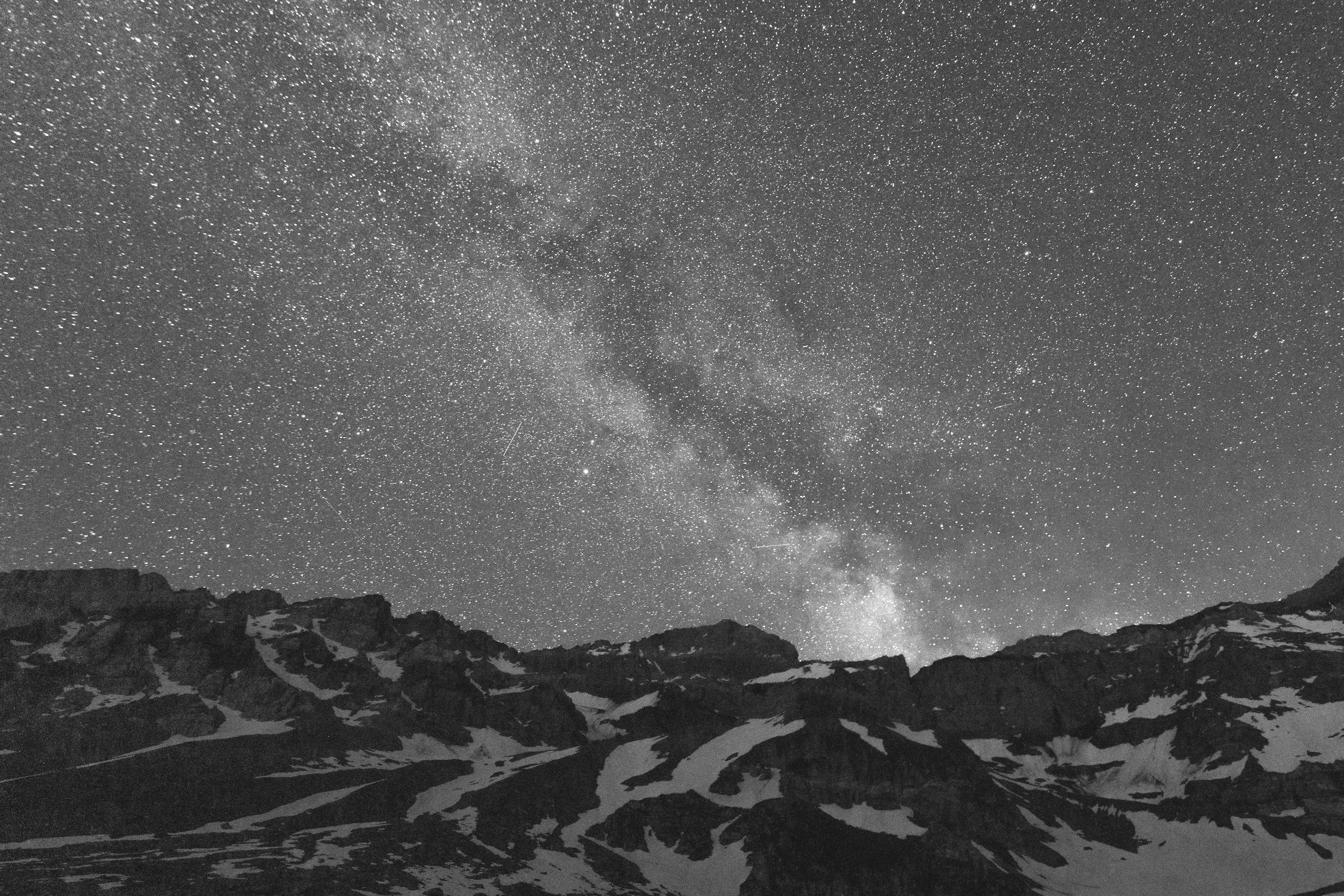 Nighttime mountain landscape with snow patches and a star-filled sky, including the Milky Way galaxy.