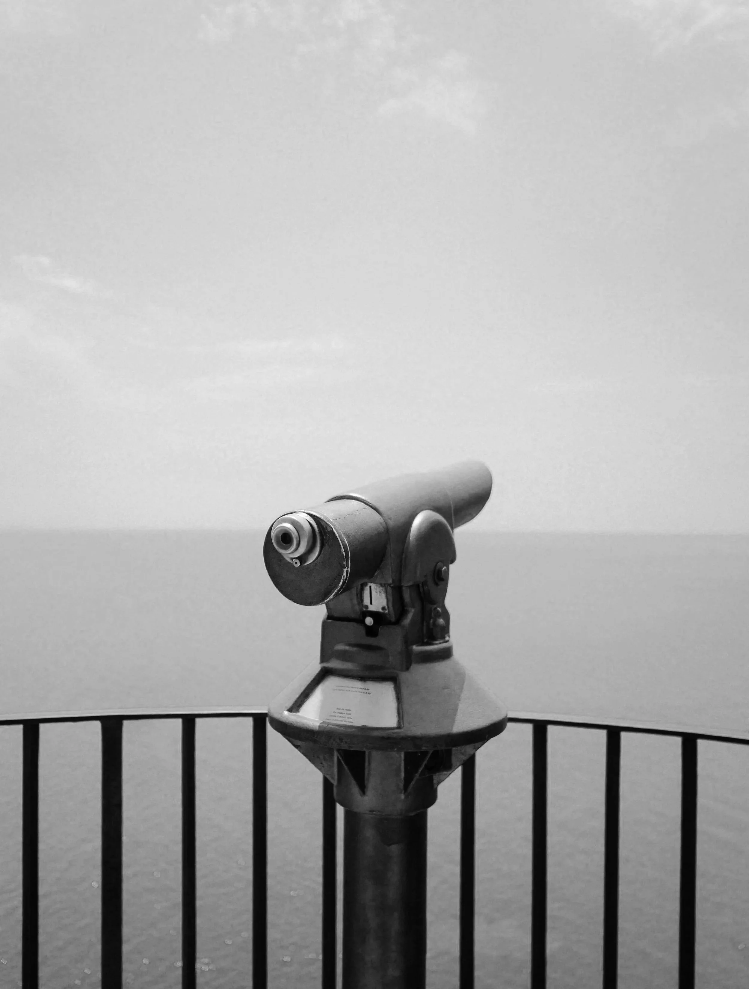 View of a coin-operated binocular viewer mounted on a stand, positioned behind a black metal fence, against a plain background.