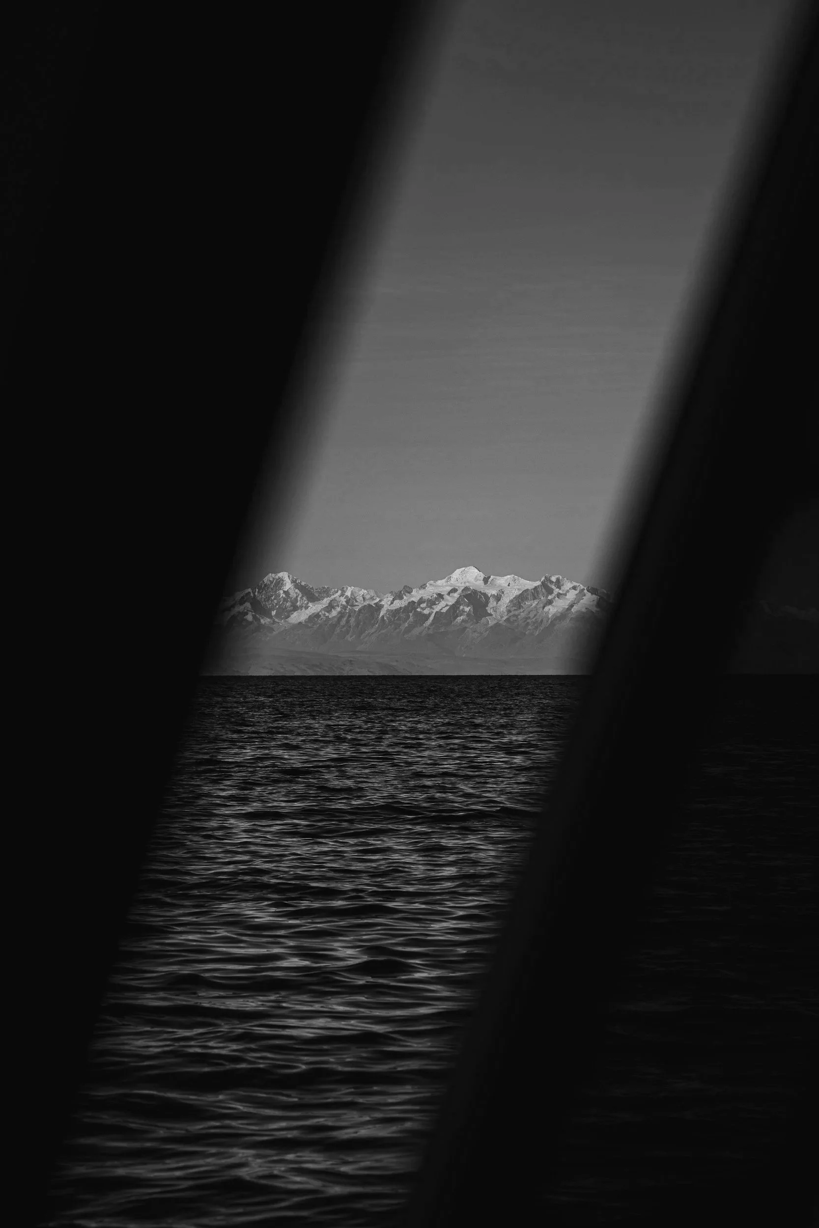 Mountain range with snow-capped peaks seen through a narrow slit or gap, with water in the foreground, in black and white.