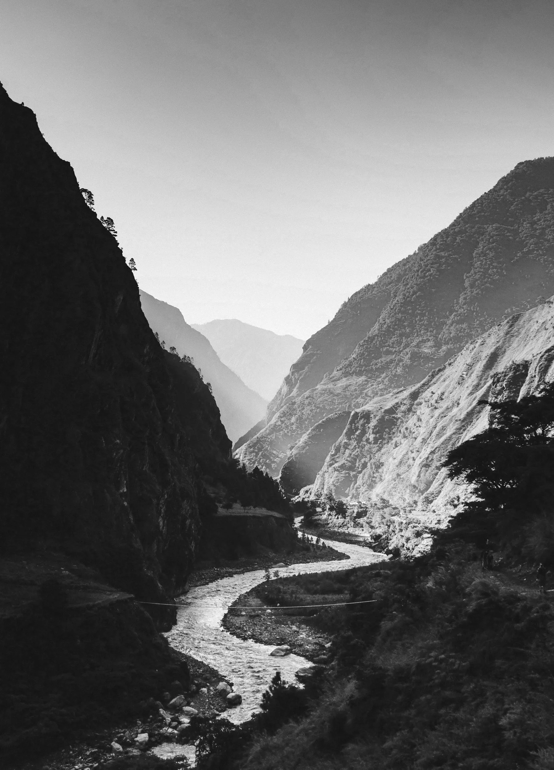 Black and white photograph of a mountain valley with a river running through it, surrounded by tall, rugged mountains and sparse trees.