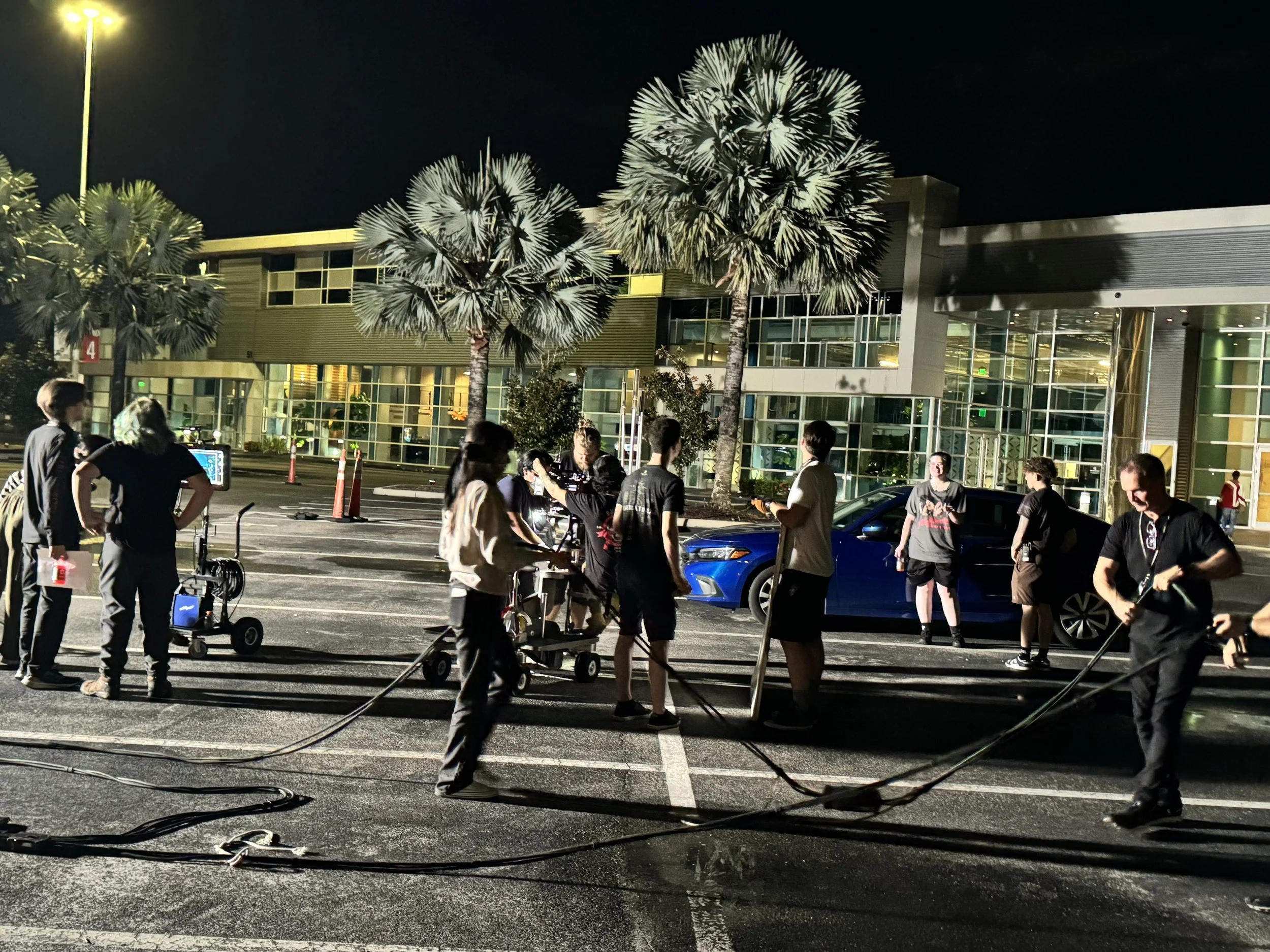 Night scene outside a modern building with illuminated windows. Several palm trees are visible, and a group of people are gathered, some with equipment and cables, possibly filming or setting up for an event.