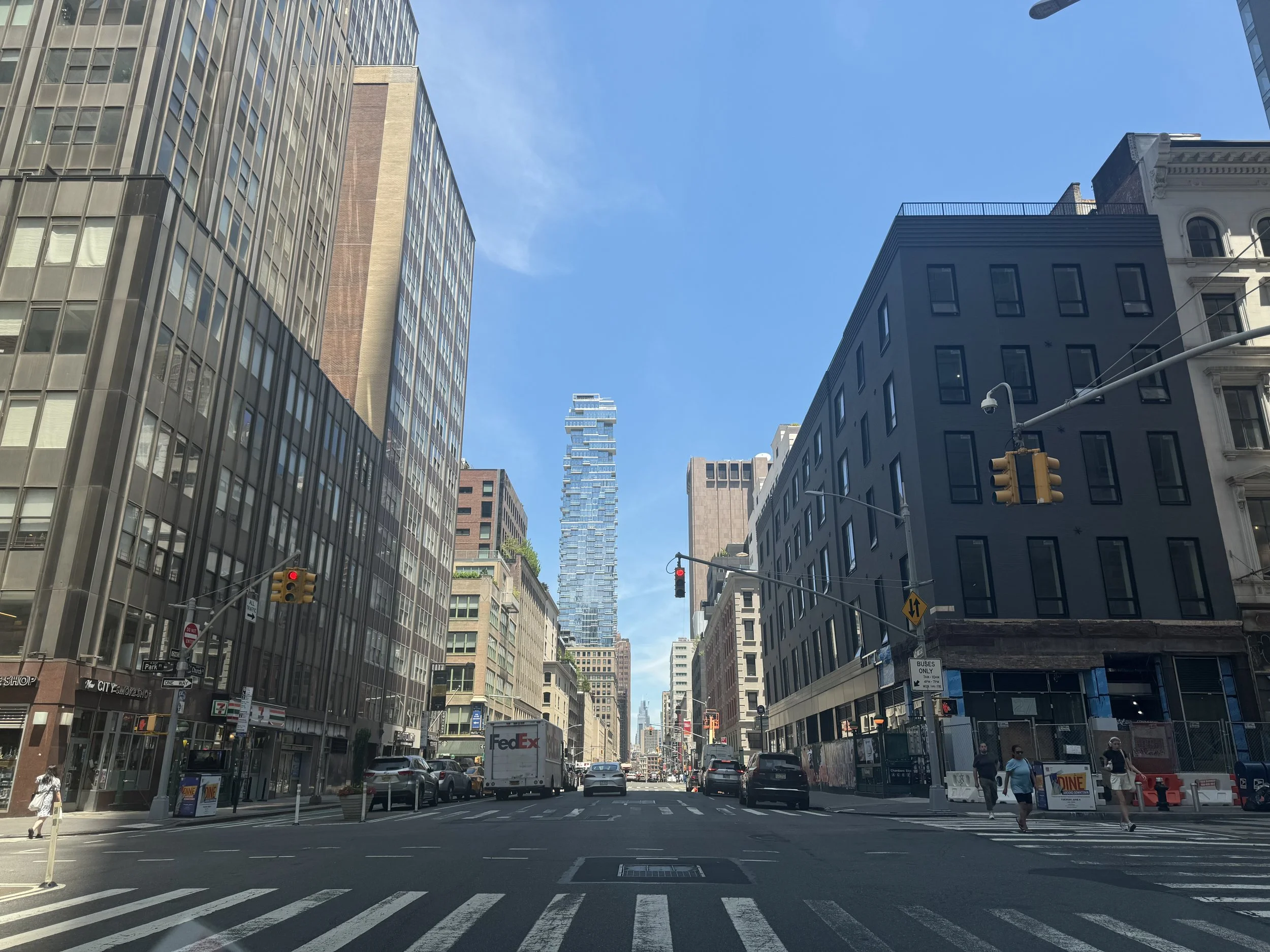 A city street scene with tall buildings on either side, cars on the road, pedestrians walking on the crosswalk, and a bright blue sky overhead.