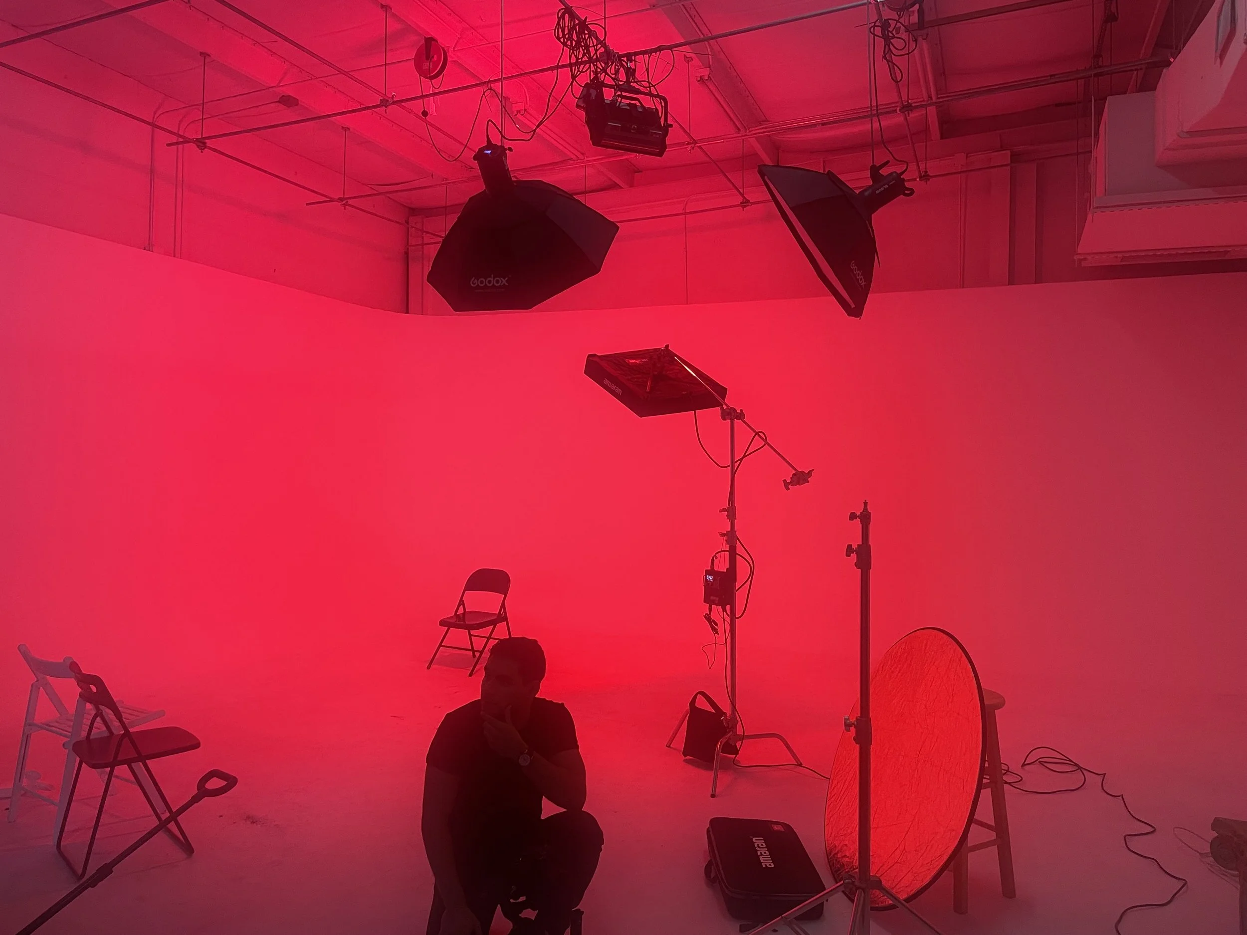 Photography studio with pink background, chairs, lighting equipment, and a person sitting in the foreground.