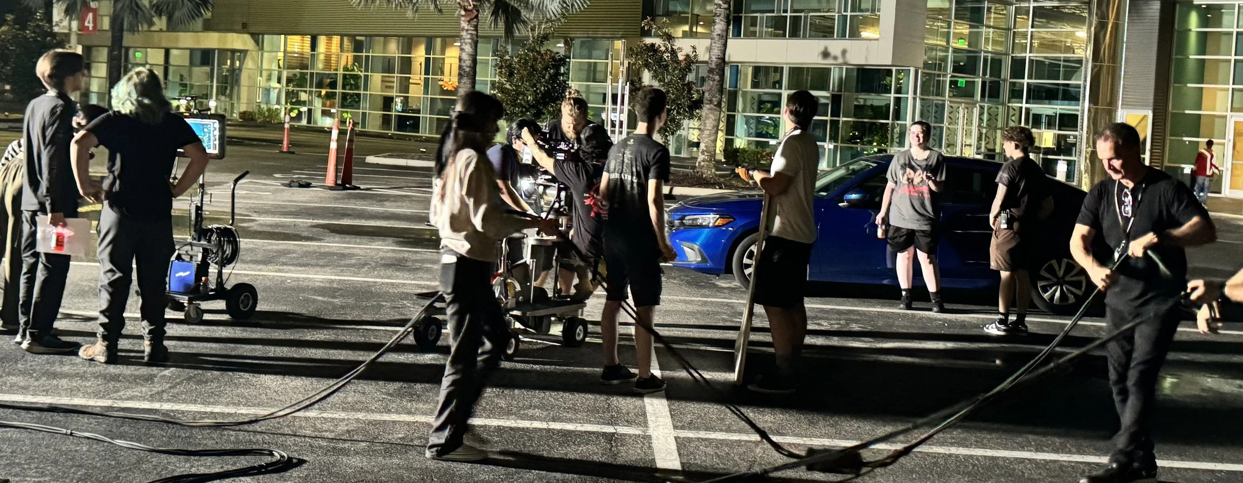 Film crew working at night in a parking lot near a modern building with glass windows, with an audience watching, film equipment, and a blue car visible.