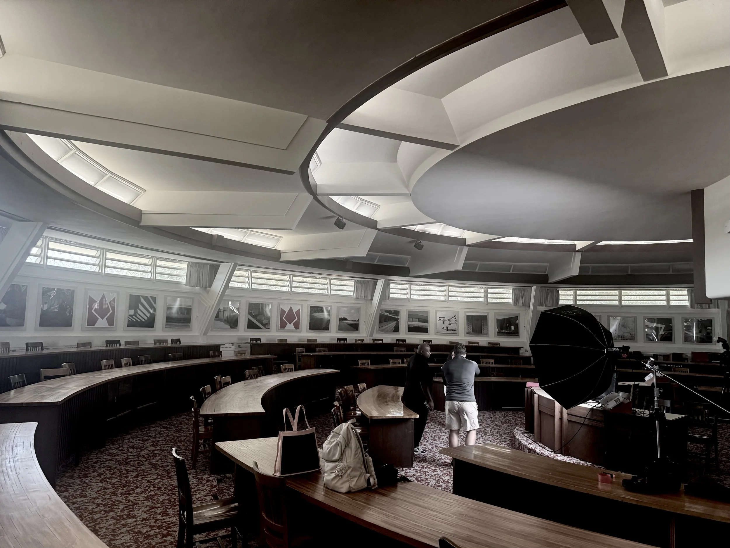Interior of a large circular room with tiered seating and a curved wooden conference table. The ceiling features a unique design with a spiral shape, and windows along the upper walls allow natural light to enter. There are two people near a grand piano on the right side of the room.