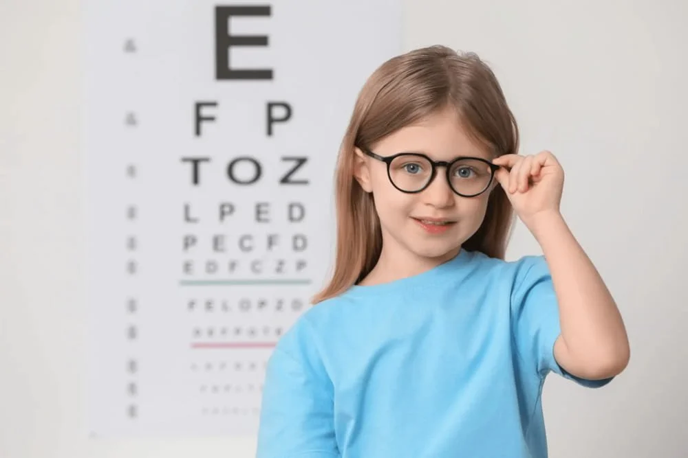 A young girl with glasses, wearing a blue shirt, standing in front of an eye chart, adjusting her glasses with her right hand.