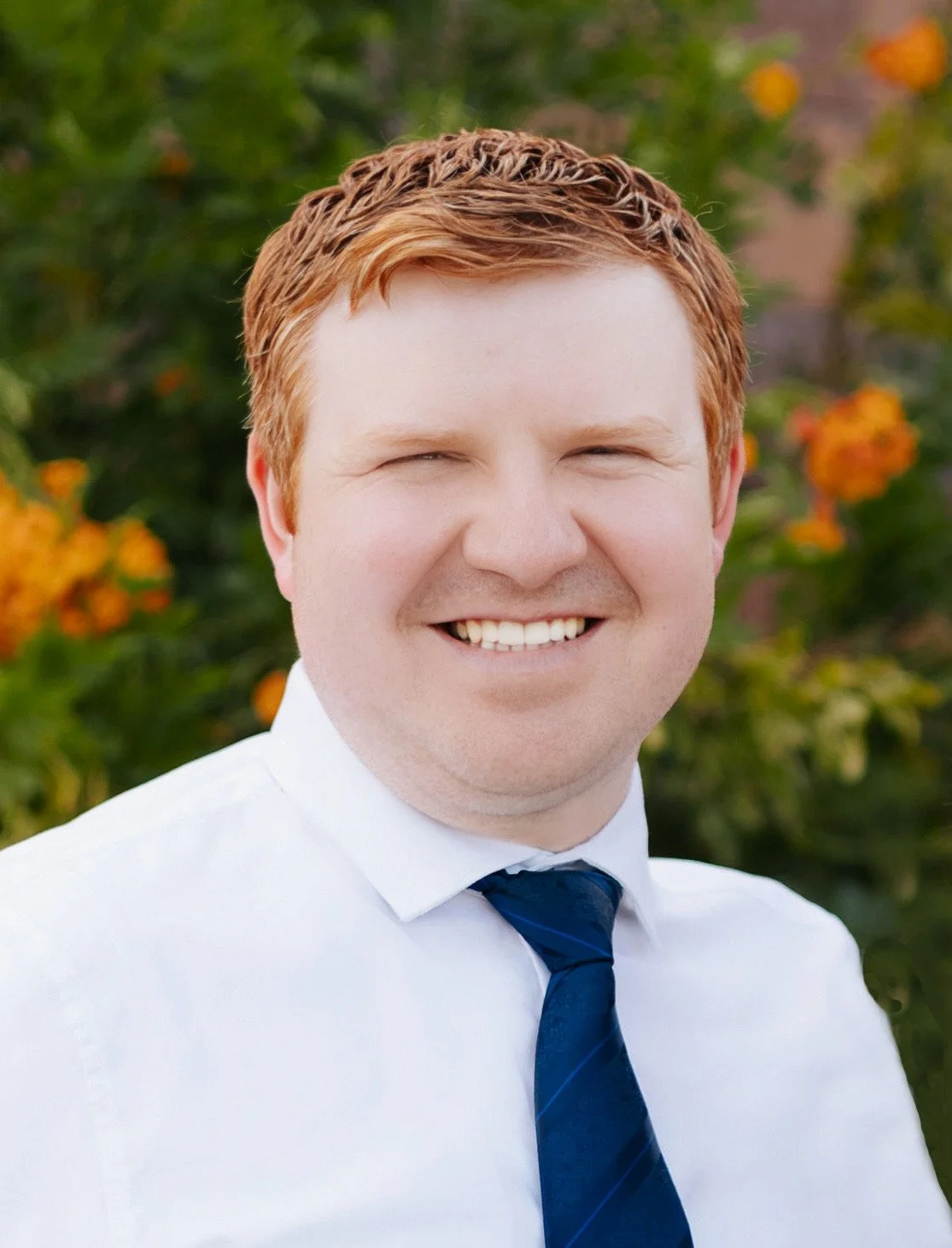 A young man with red hair, smiling, wearing a white shirt and a dark blue tie, standing outdoors with orange flowers and green foliage in the background.