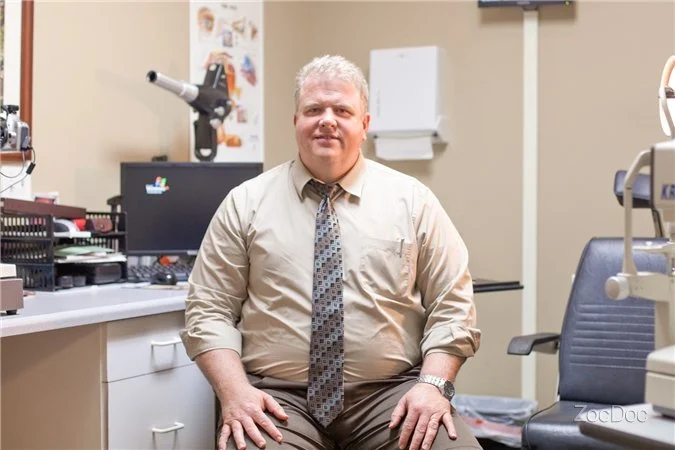 A man sitting in an optometry office. He is wearing a beige shirt and tie, seated in front of equipment and a computer. The office has medical instruments and charts visible in the background.