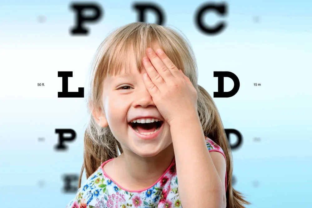 A young girl laughing with her hand covering one eye. She is in front of a blurred eye chart background.