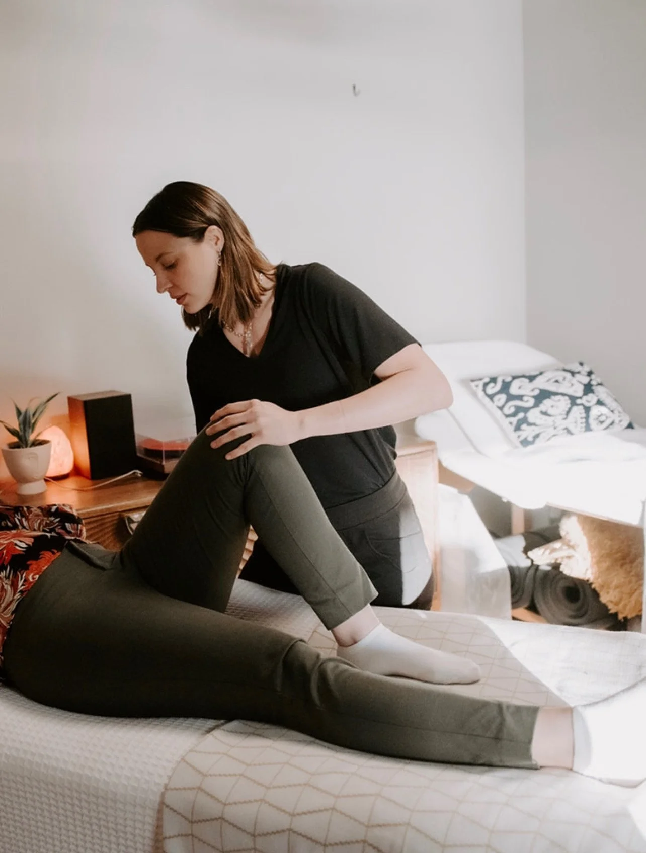 A woman with shoulder-length brown hair, wearing a black t-shirt and gray pants, assisting a person in a black floral shirt and pants, lying on a bed, in a bedroom with a wooden nightstand and decorative elements.