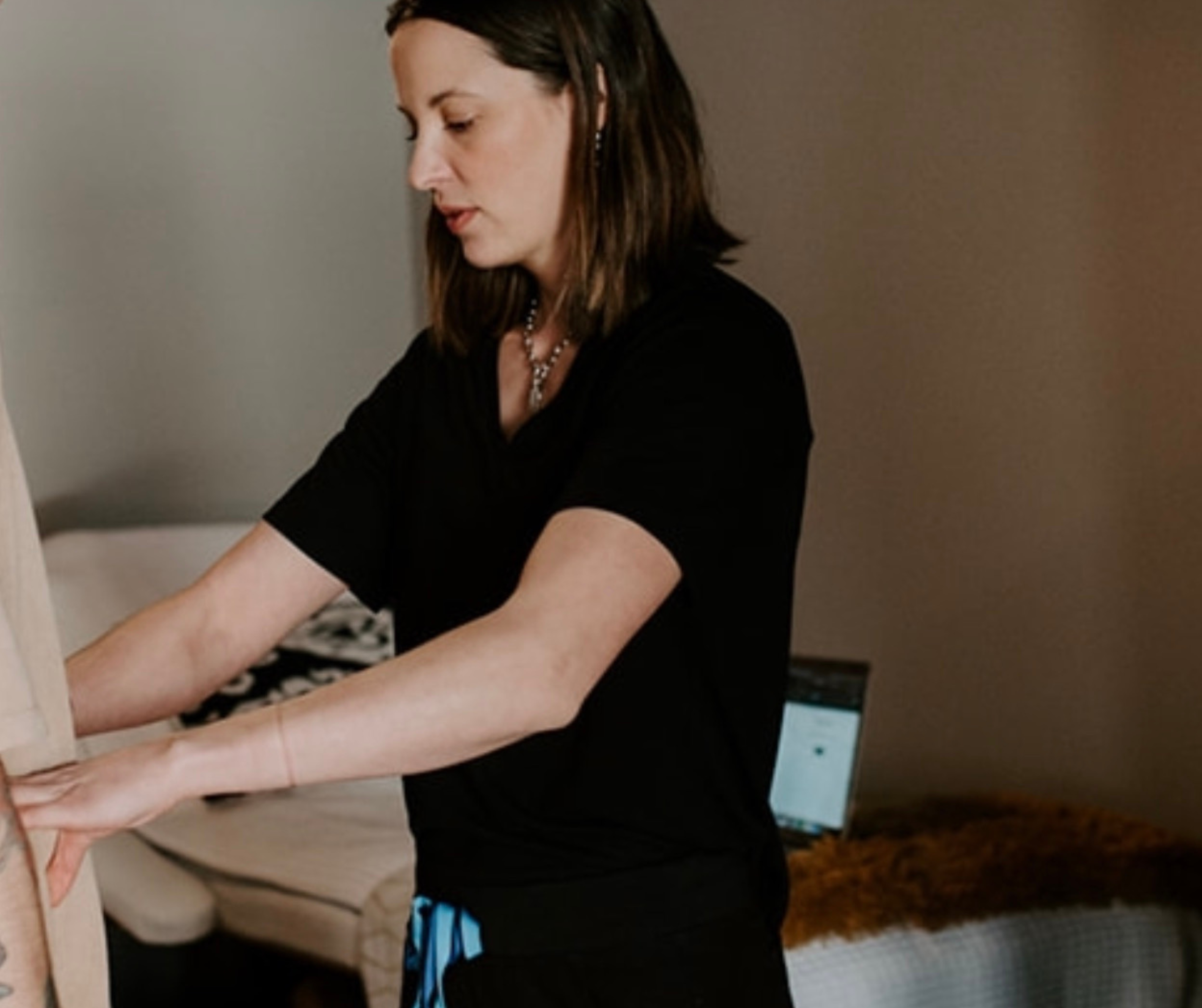A woman with shoulder-length brown hair, wearing a black shirt and jewelry, standing indoors with her hands on a wooden object, with a bed and a laptop in the background.