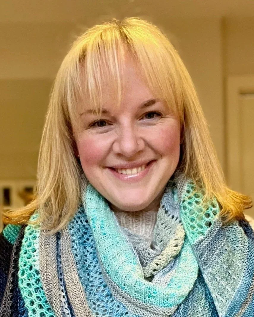 A blonde woman smiles at the camera. She is wearing a blue, hand knitted scarf