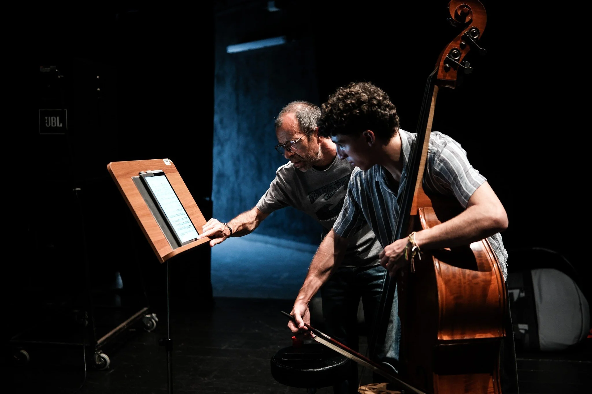 Duas pessoas, uma mais velha e uma mais jovem, observando uma partitura em uma tela digital enquanto praticam música com um violoncelo, em um ambiente escuro.
