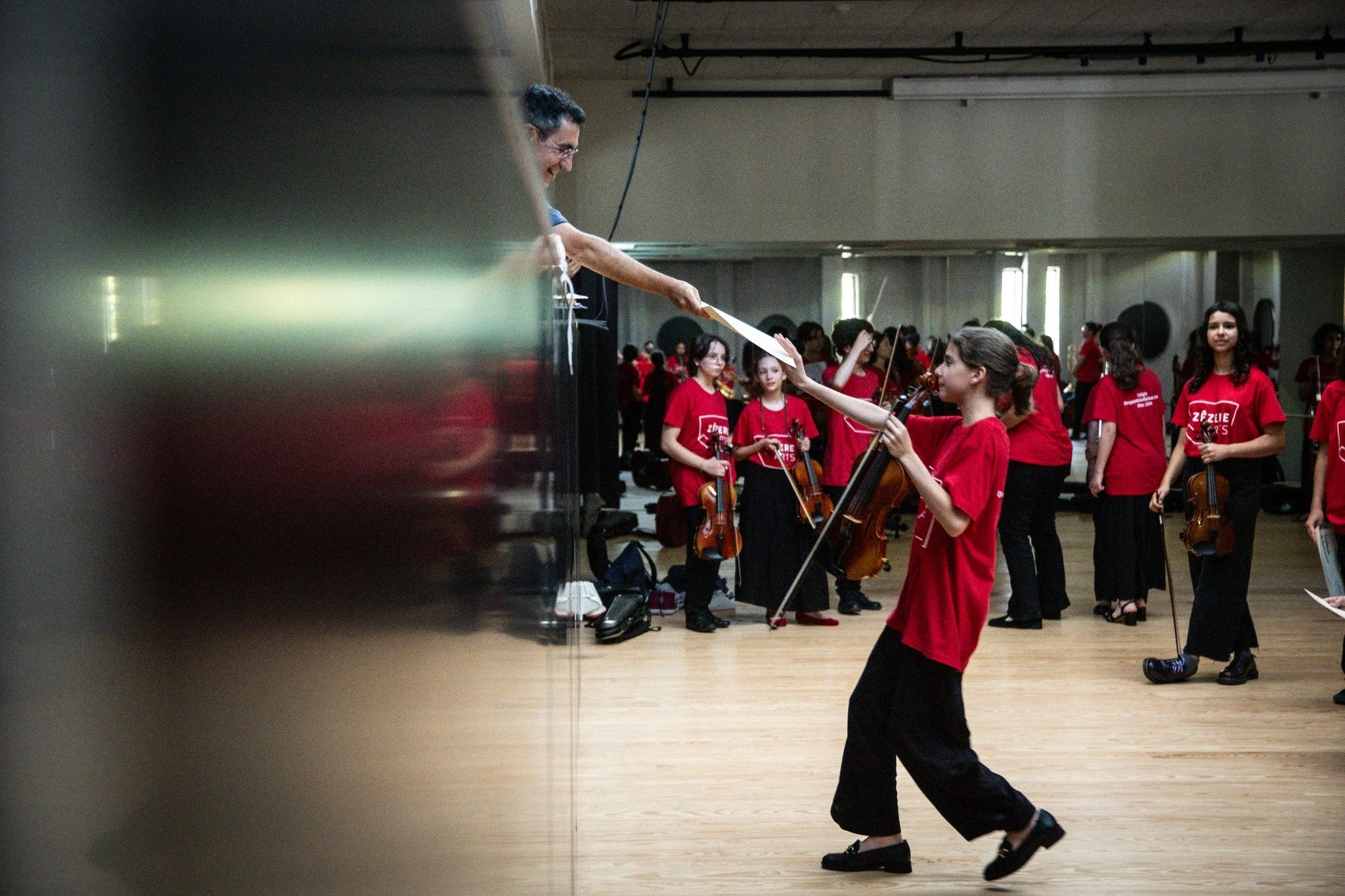 Mulher entregando uma partitura para uma jovem violinista durante ensaio de orquestra com várias crianças tocando violino, todas usando camiseta vermelha, em uma sala de prática.