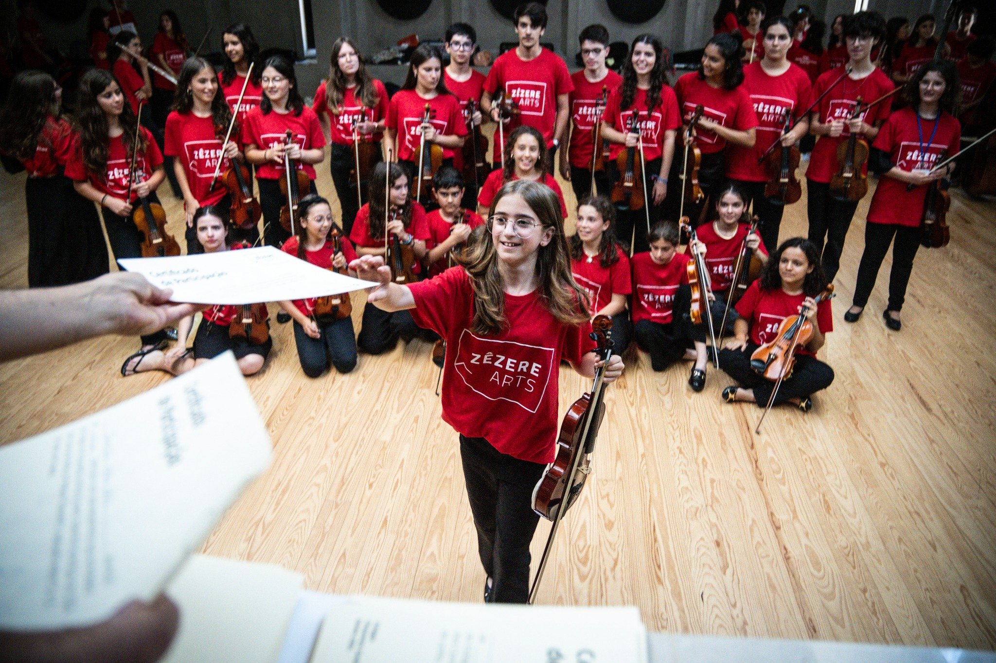 Um grupo de crianças e adolescentes com camisetas vermelhas do evento 'Zézere Arts' posando para a foto, segurando violinos, durante uma apresentação ou ensaio, em um ambiente interno com piso de madeira.