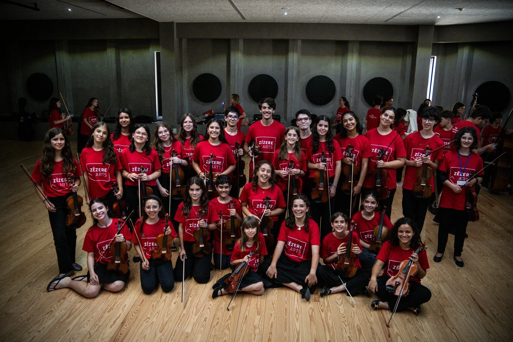 Grupo de jovens músicos de orquestra com instrumentos de corda, todos usando camisetas vermelhas com a escrita 'Zézère Arts'.