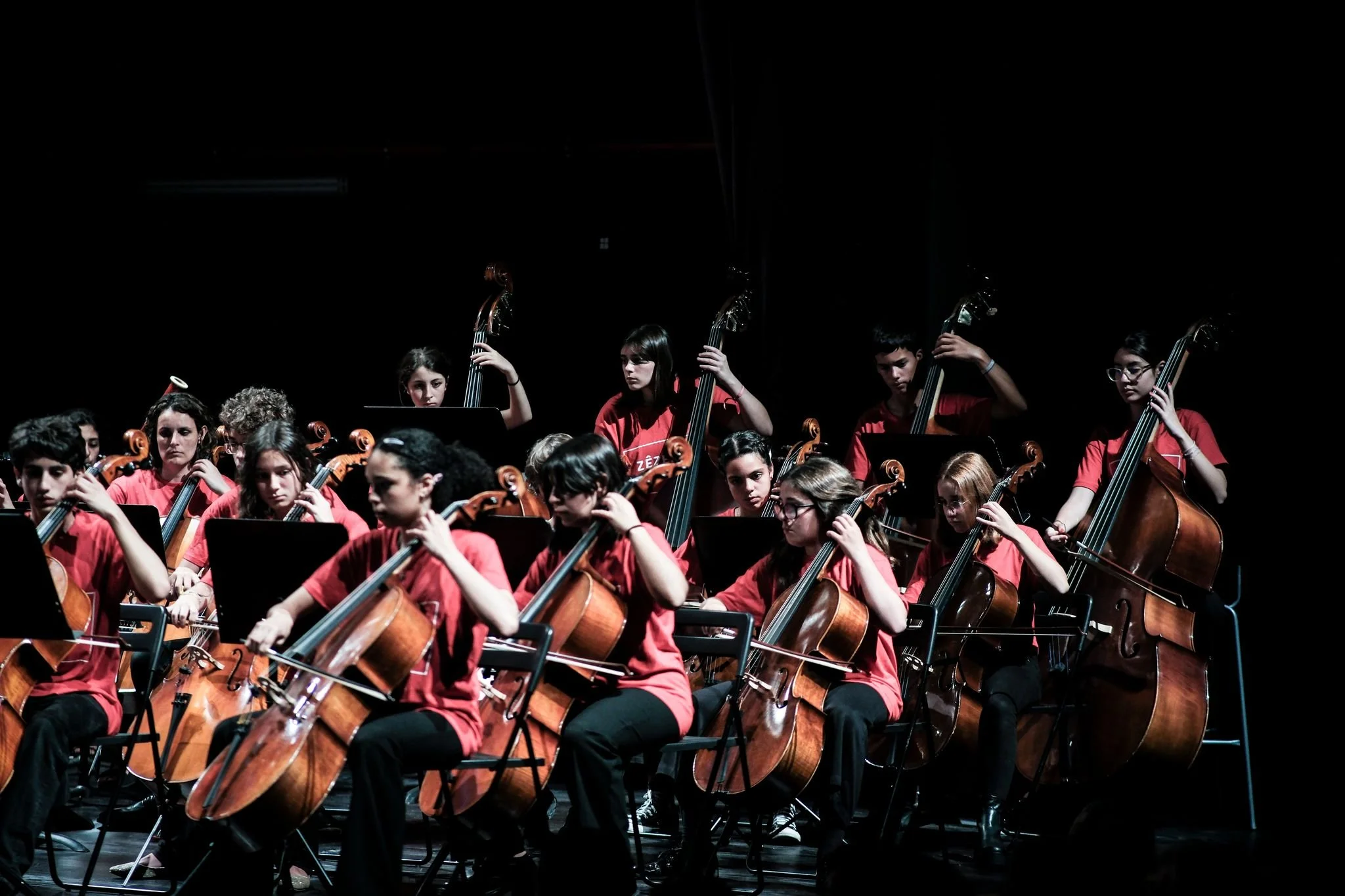 Orquestra de jovens músicos tocando violoncelos e baixos em palco escuro, todos de camiseta vermelha.