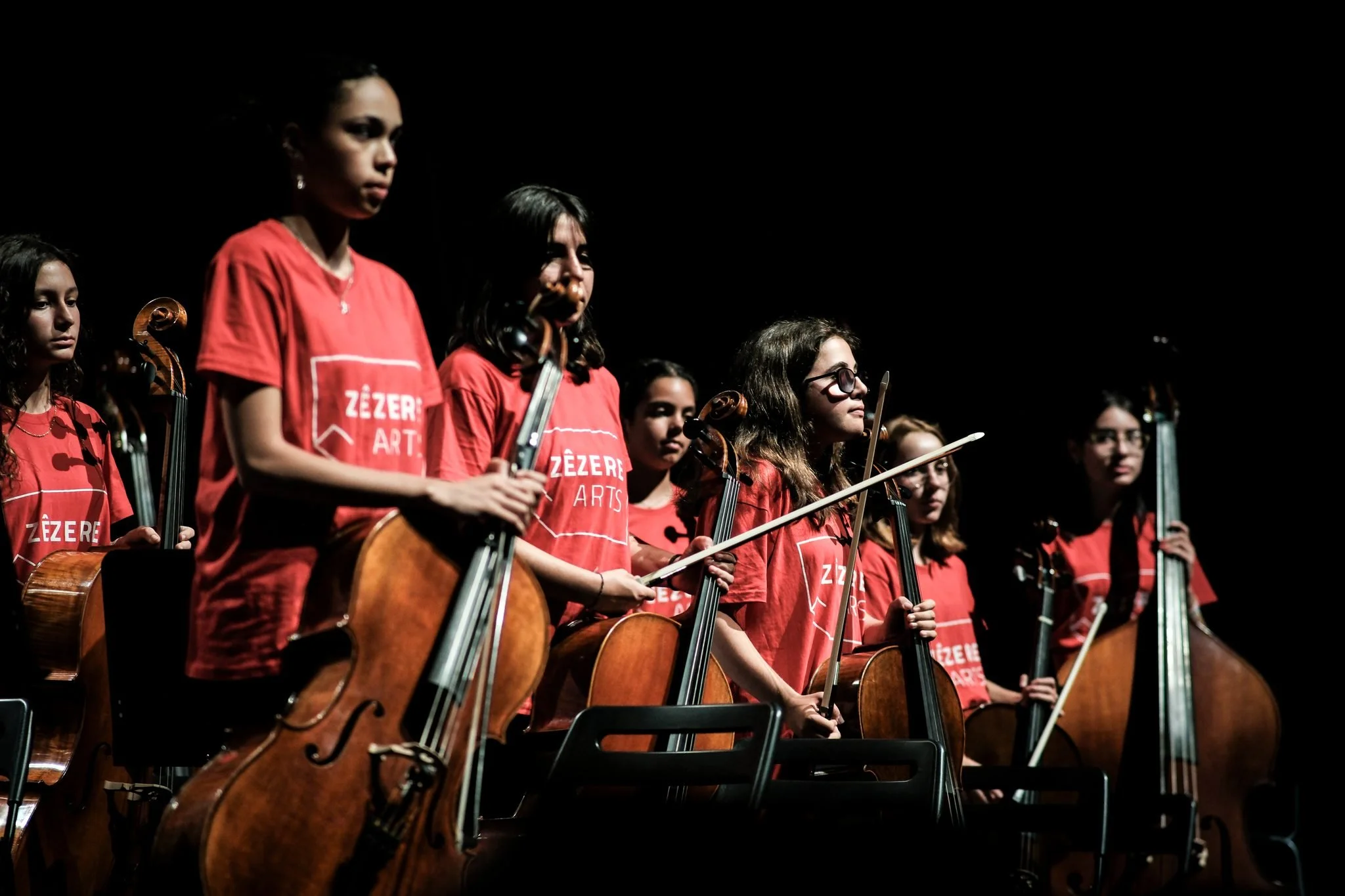 Grupo de jovens musiciansas tocando violoncelo, algumas usando camisetas vermelhas com o logo 'Zêzere Arts' em um palco.
