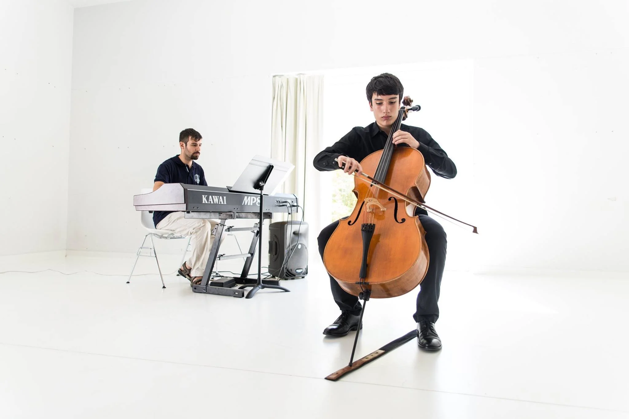 Dois músicos tocando instrumentos musicais em uma sala branca: um tocando violoncelo e outro tocando teclado
