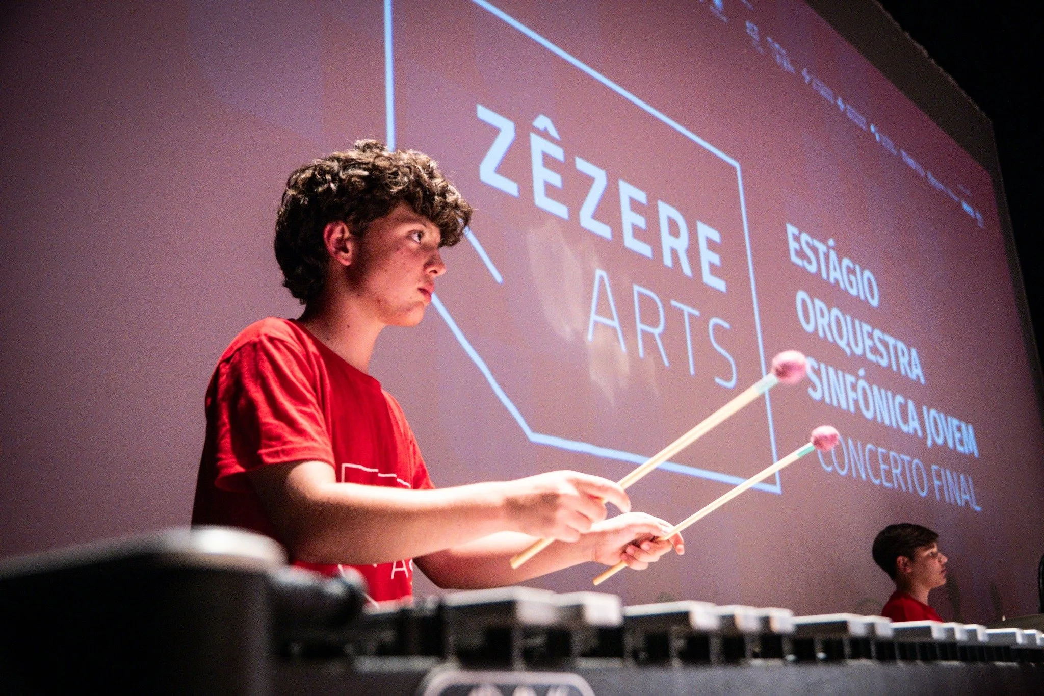Jovem tocando um instrumento musical em uma apresentação com uma tela ao fundo exibindo o nome 'Zêzere Arts' e uma lista de atividades como estágio, orquestra, sinfônica jovem e concerto final.