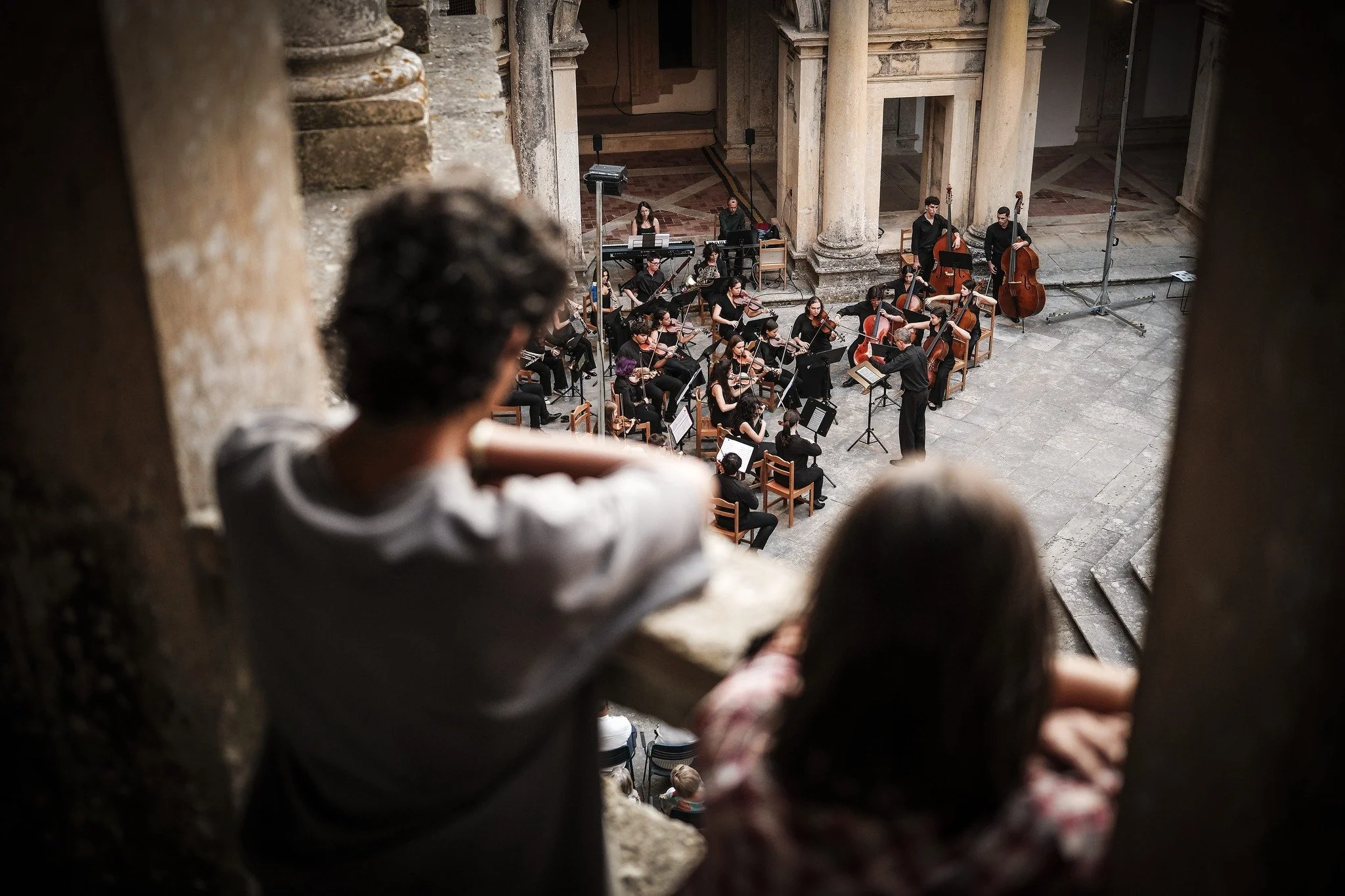 Uma orquestra se apresentando em um palco de uma sala de concertos com paredes de pedra e colunas. Dois espectadores, um menino e uma menina, observam a apresentação de uma varanda, com o menino de cabelo escuro e a menina com cabelo castanho, ambos 