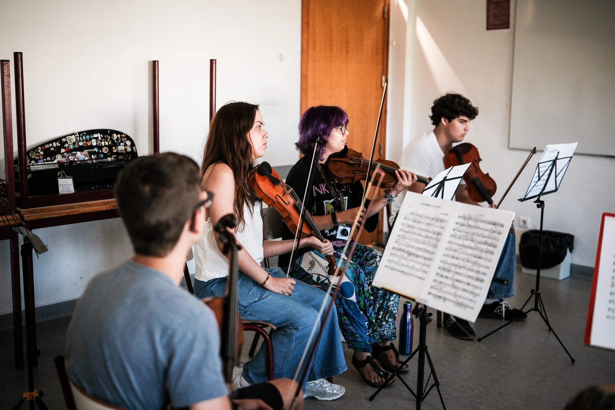 Grupo de jovens músicos tocando violino em uma sala de ensaio com partituras e instrumentos musicais ao fundo.